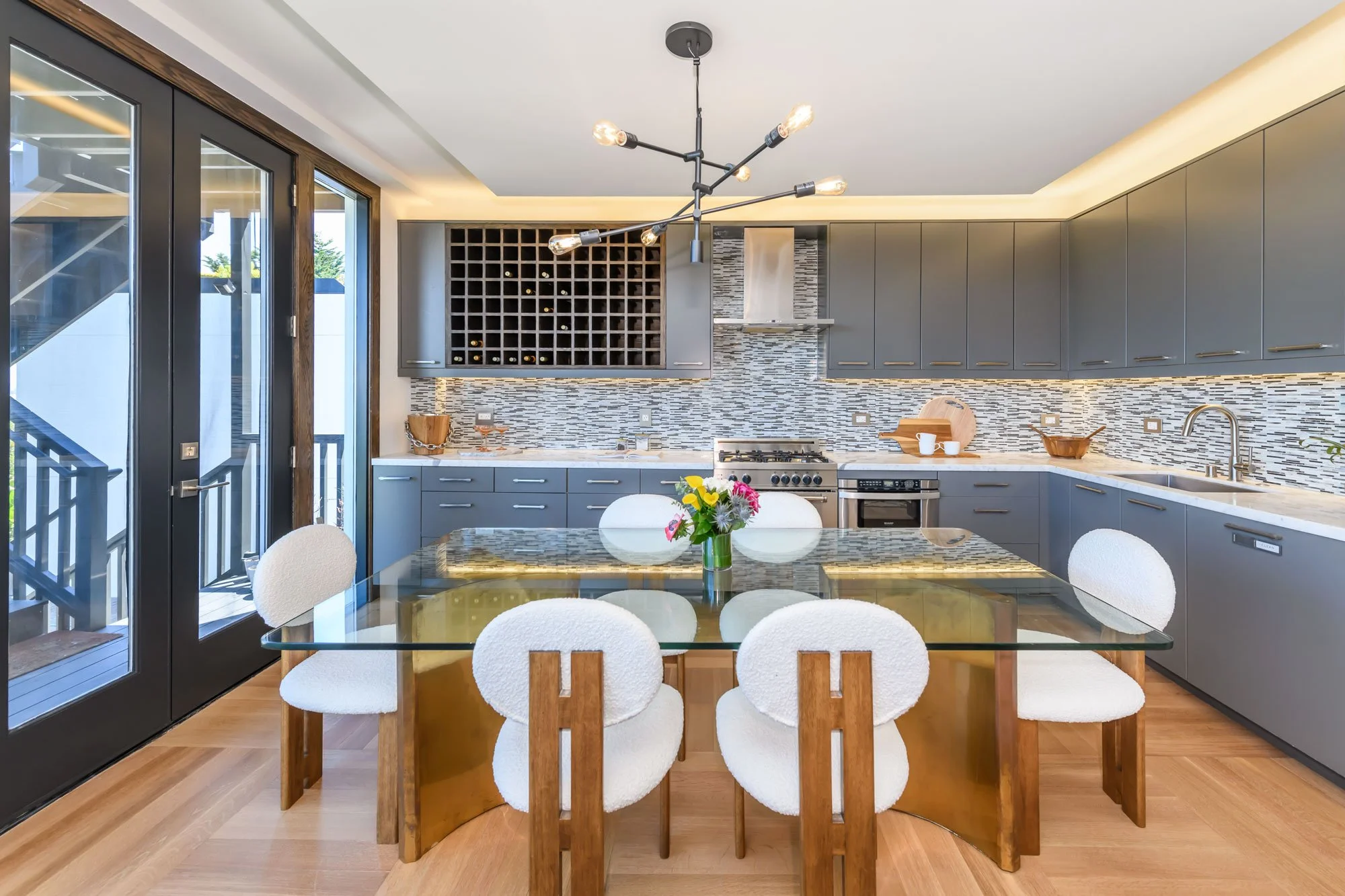 Modern kitchen with gray cabinets, a mosaic tile backsplash, a wine rack, a stainless steel range, and a glass-top dining table with white upholstered chairs.