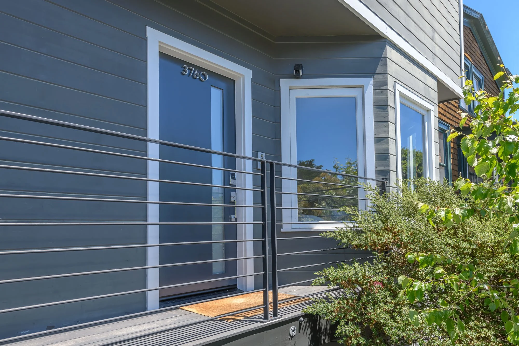 Close-up of a modern house facade with gray and wooden siding, large windows, and a balcony with a metal railing, surrounded by trees and greenery.