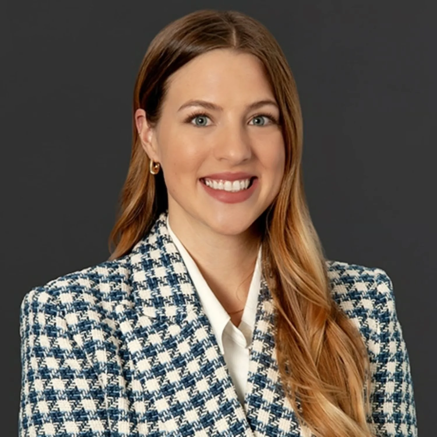 Portrait of a woman with long red hair, wearing a checkered blazer and white shirt, smiling, against a dark gray background.