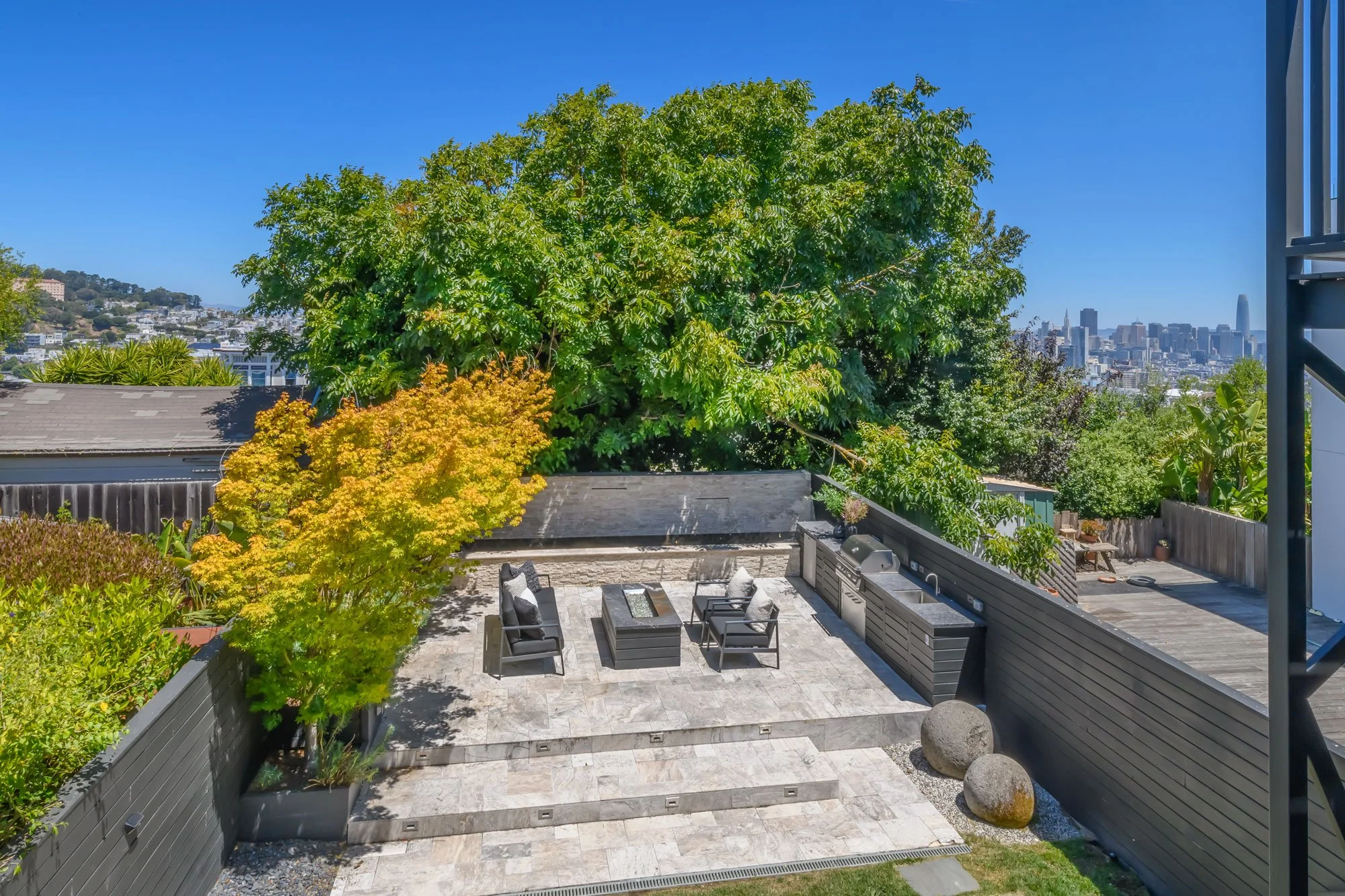 A spacious outdoor patio with modern furniture, surrounded by lush greenery and city skyline in the background under clear blue skies.