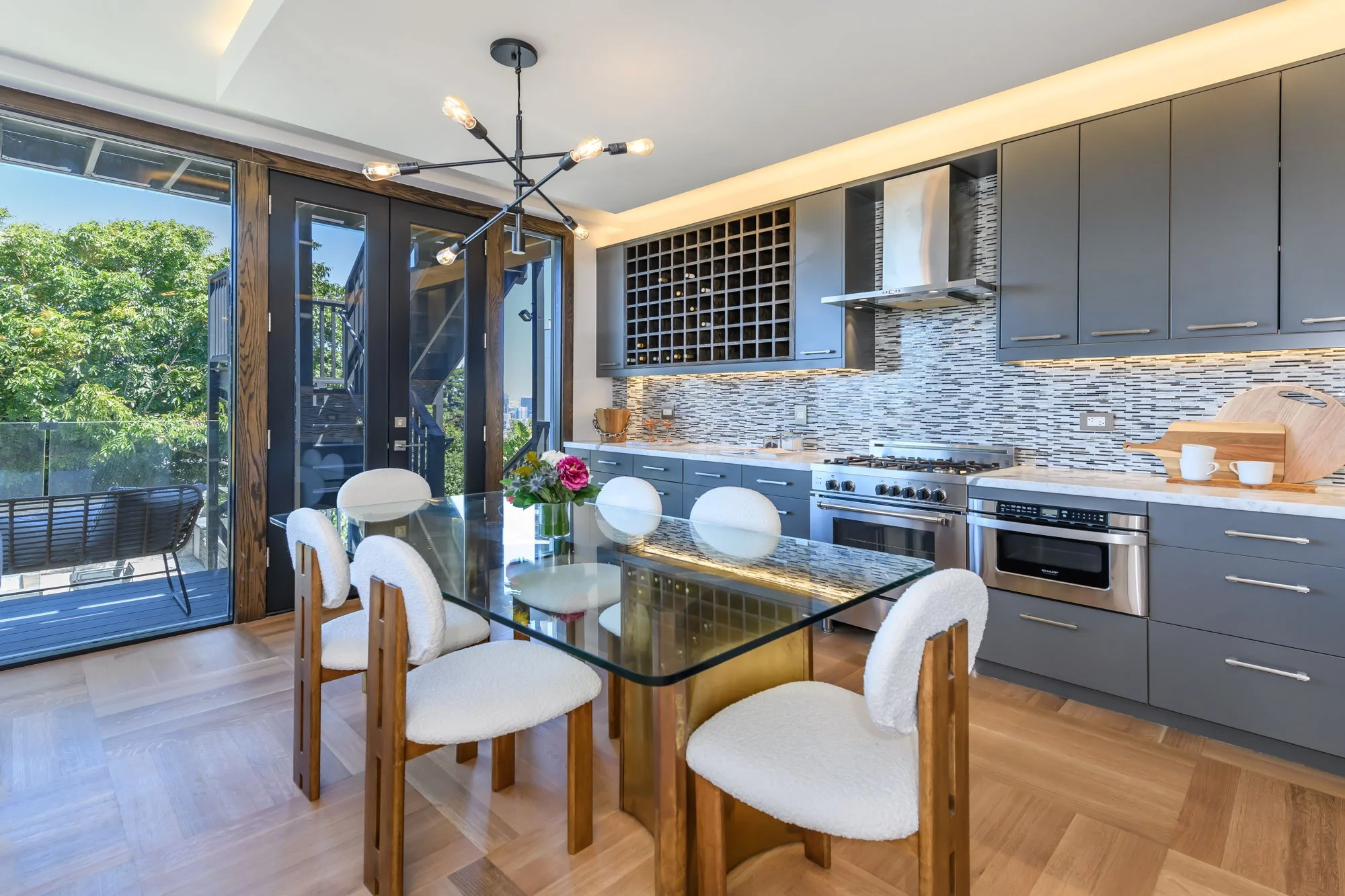 Modern kitchen with gray cabinets, mosaic backsplash, stainless steel appliances, and a glass dining table with white chairs. Large glass doors open to a balcony with outdoor seating and greenery.