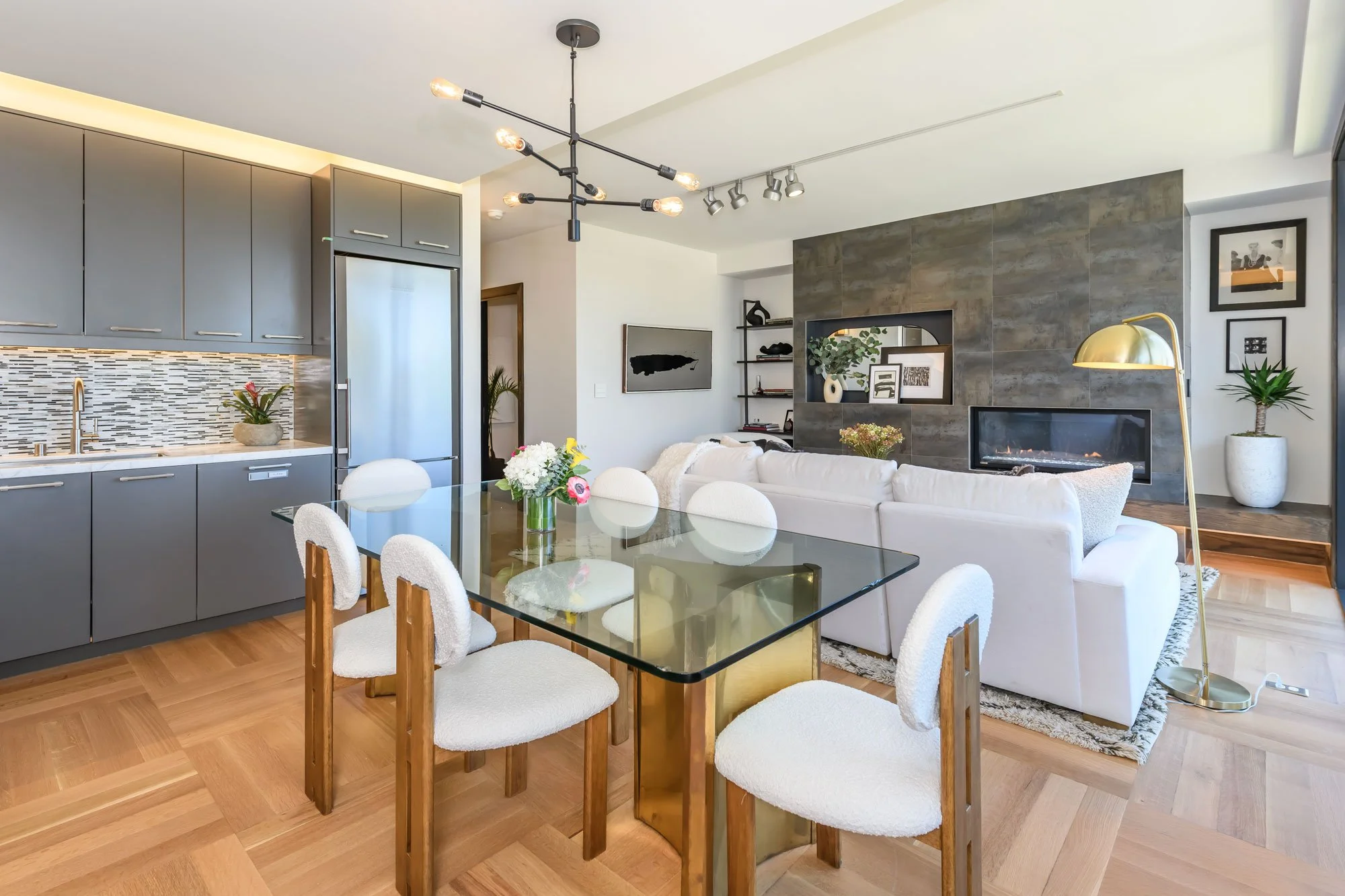 Open-concept living and dining area with gray kitchen cabinets, a glass dining table with white upholstered chairs, a white sectional sofa, a modern chandelier, a fireplace, and decorative plants and artwork.