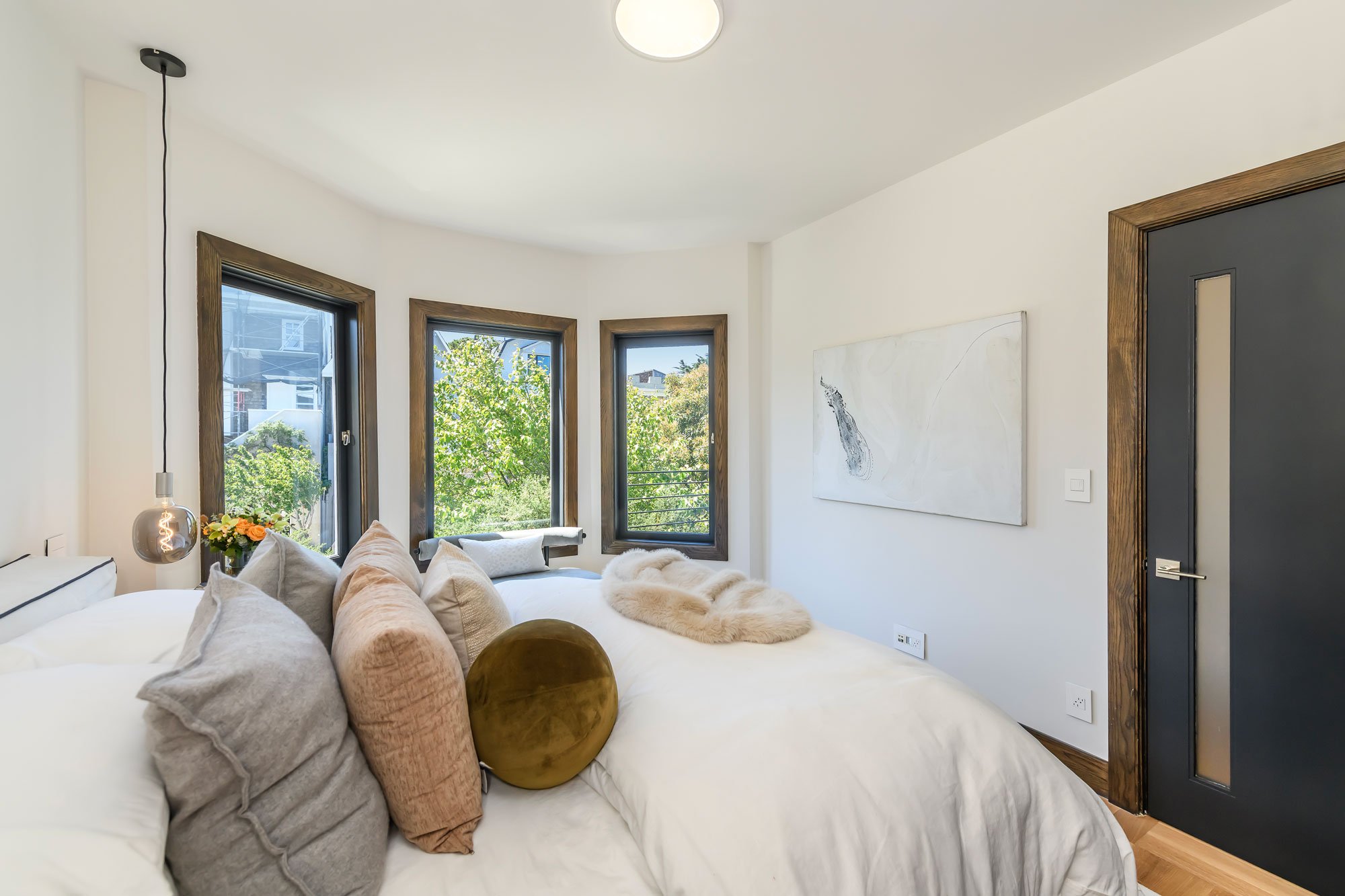 A cozy living room with a white sofa decorated with beige, gray, and velvet pillows, a furry throw blanket, and a black window with wooden trim showing green trees outside. There's a modern pendant light hanging from the ceiling, a large abstract pai