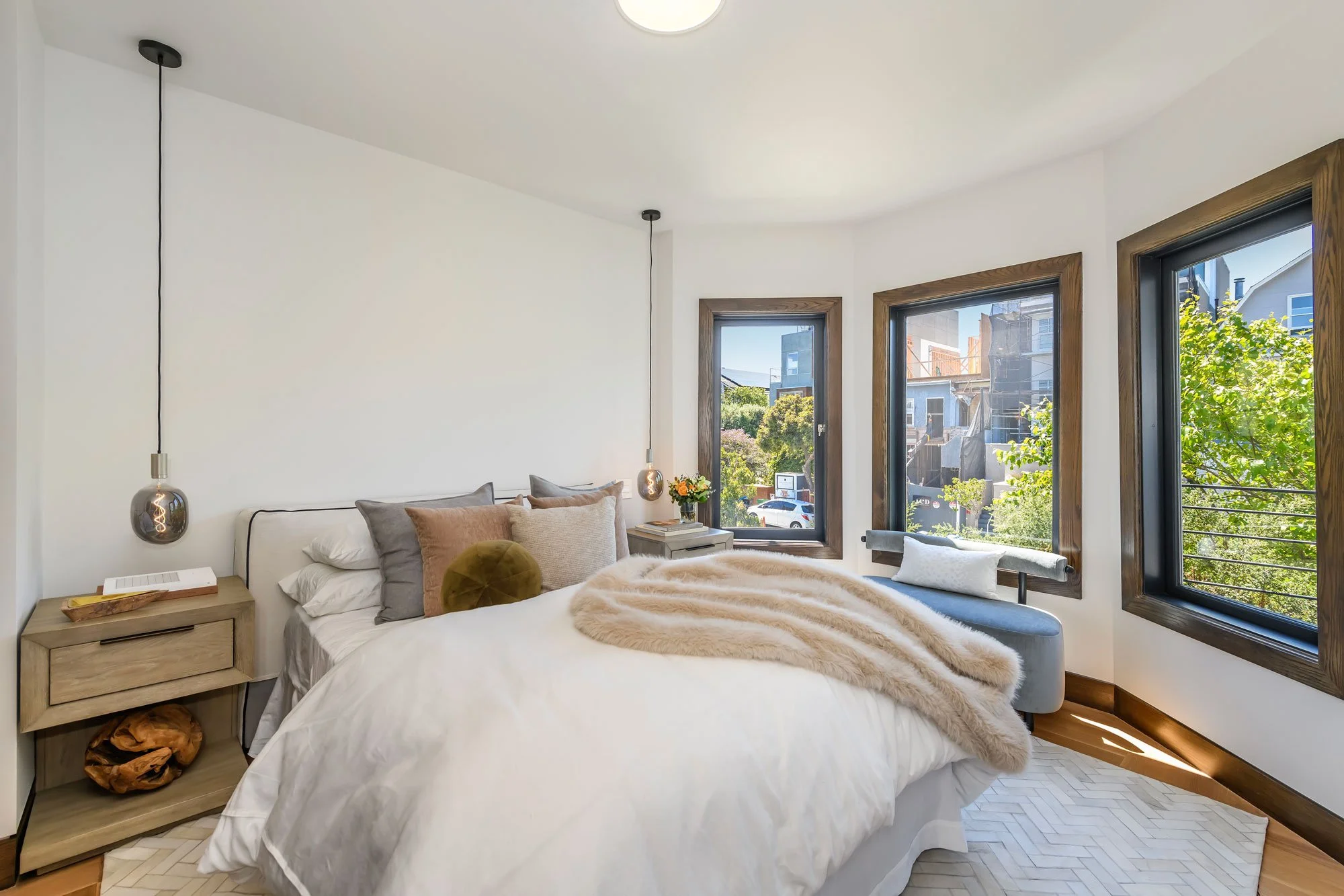 Modern bedroom with white walls and large windows showing neighboring houses and trees outside. The bed has multiple pillows and a beige blanket. There is a bedside table with books and a decorative item. Two hanging pendant lights are above the bed,