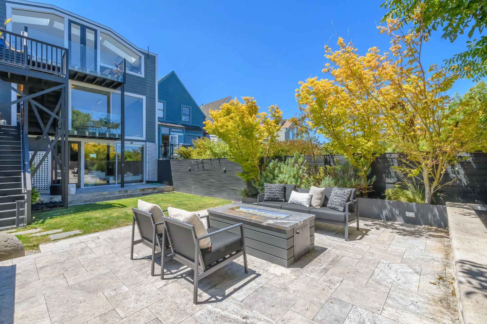 Modern backyard with outdoor seating area featuring a fireplace, trees with green and yellow leaves, and contemporary house with large windows and multiple balconies.