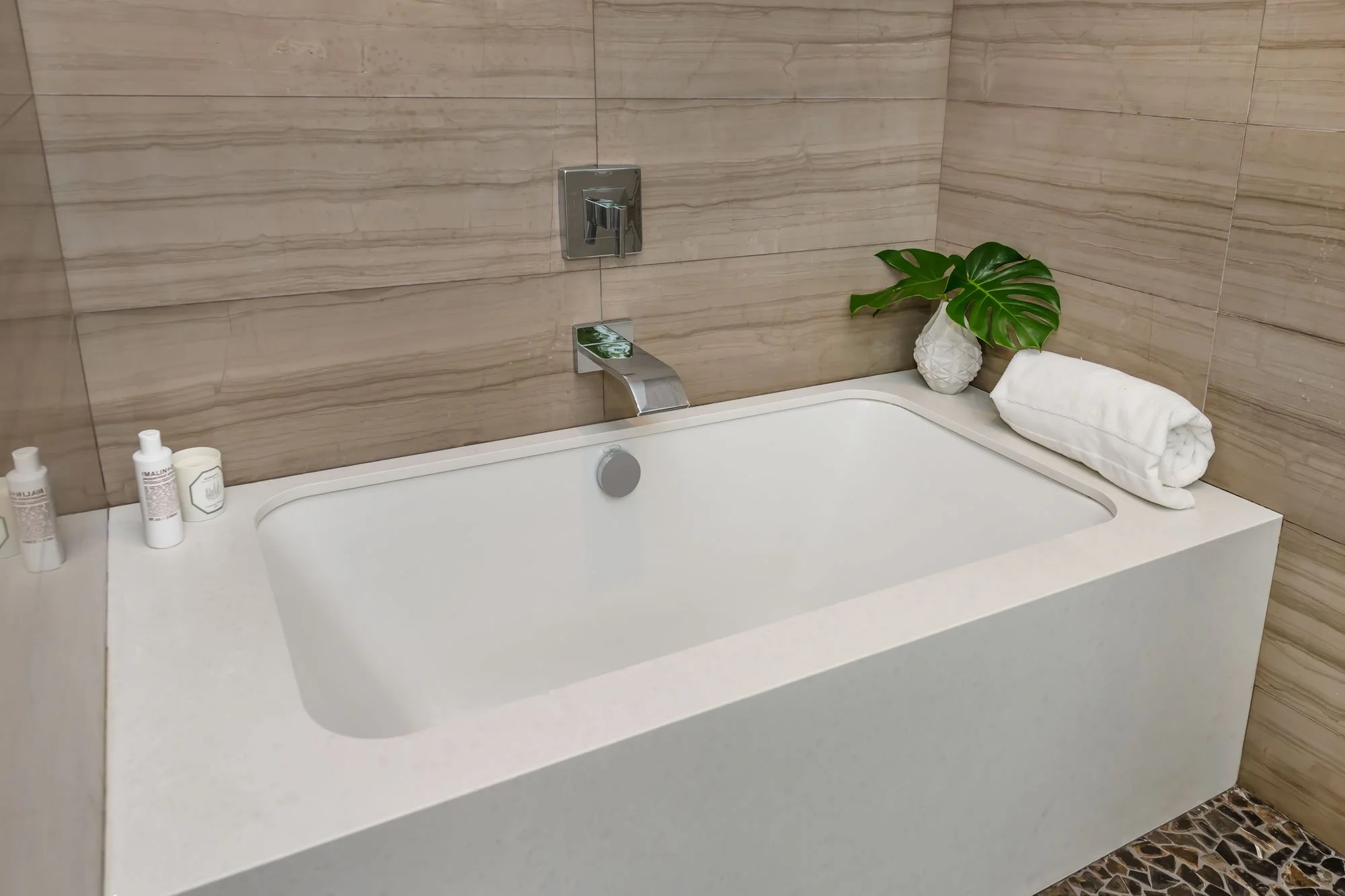 A bathroom with beige tiled walls, a white bathtub, a chrome faucet, and a small potted monstera plant with green leaves placed on the edge of the tub. There is a rolled white towel next to the plant, and bottles of bath products on the side of the t