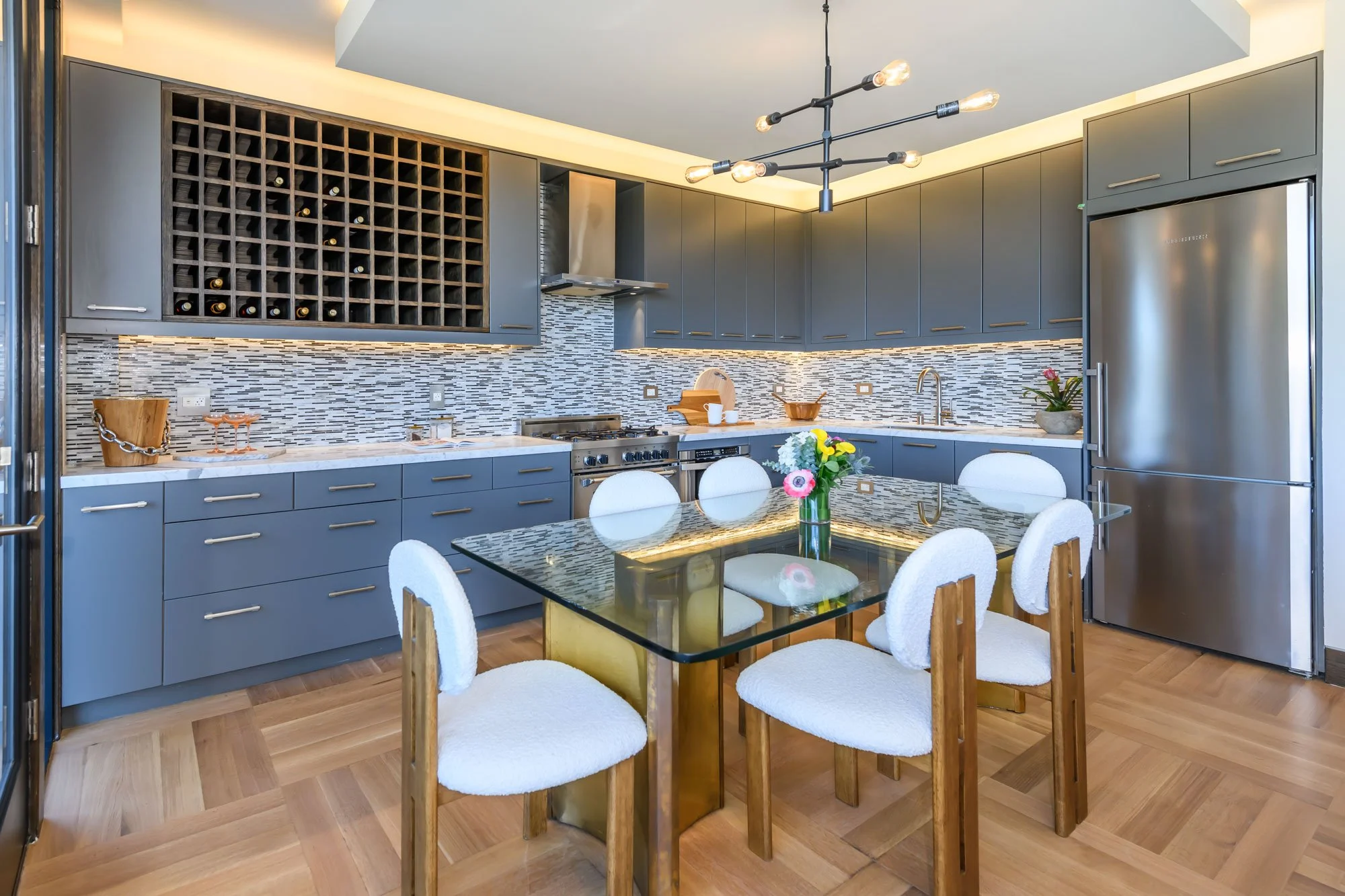A modern kitchen with blue cabinets, a mosaic tile backsplash, and a glass-top dining table surrounded by white upholstered chairs. There is a wine rack above the counter, a stainless steel refrigerator, and a chandelier with exposed bulbs hanging fr