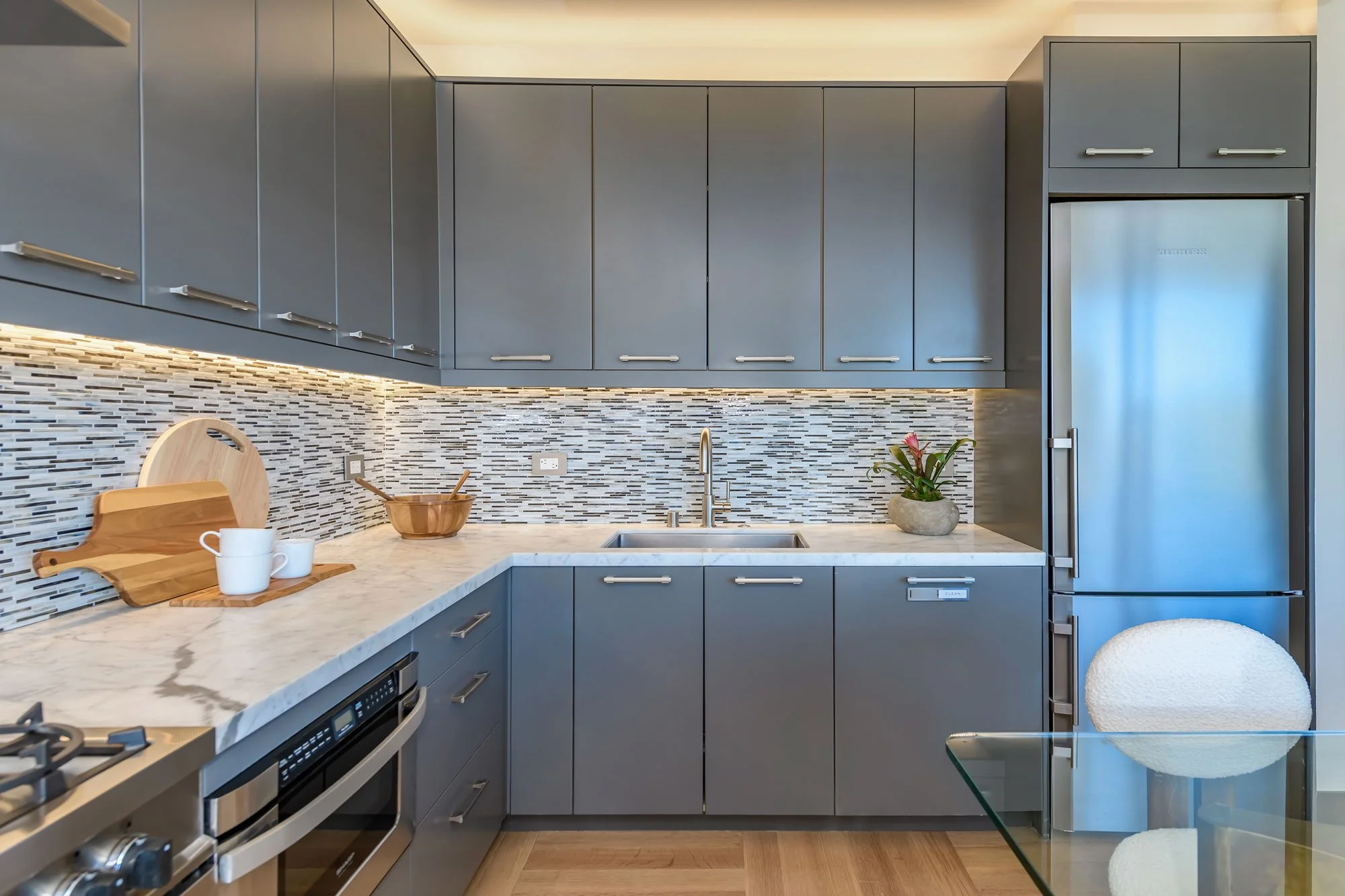 Modern kitchen with blue cabinets, white marble countertop, and a mosaic tile backsplash. Contains a stainless steel refrigerator, a gold sink, a wooden cutting board, and white mugs on the counter. There are also plants and a glass table with a roun