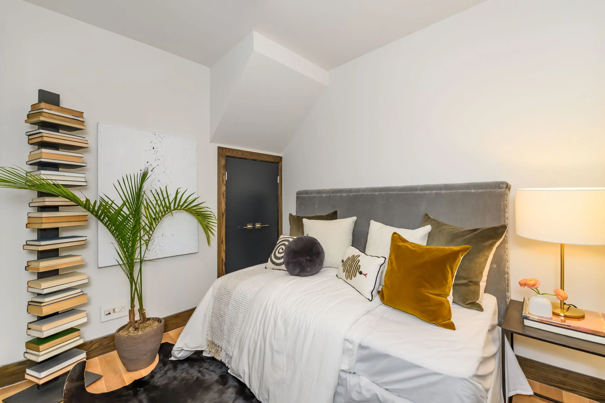 A modern bedroom with a large bed, multiple pillows, and a gray headboard. To the right, there is a bedside table with a lamp and decor. On the left, a tall, vertical stack of books is mounted on the wall beside a potted plant. There is a dark door w