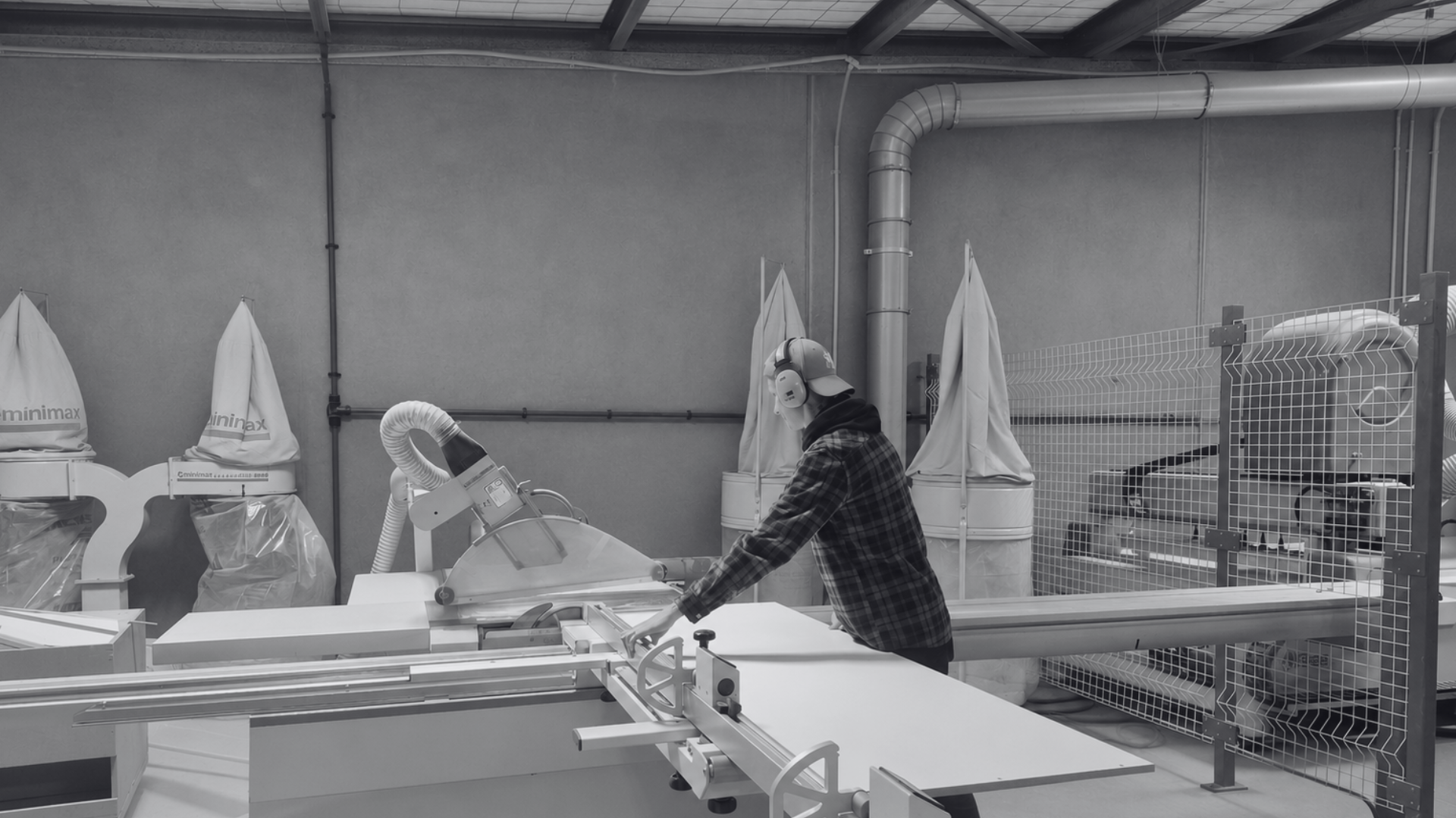 A worker in a plaid shirt and headphones operating a cutting machine in an industrial setting, with large fabric hoods and ventilation pipes in the background.