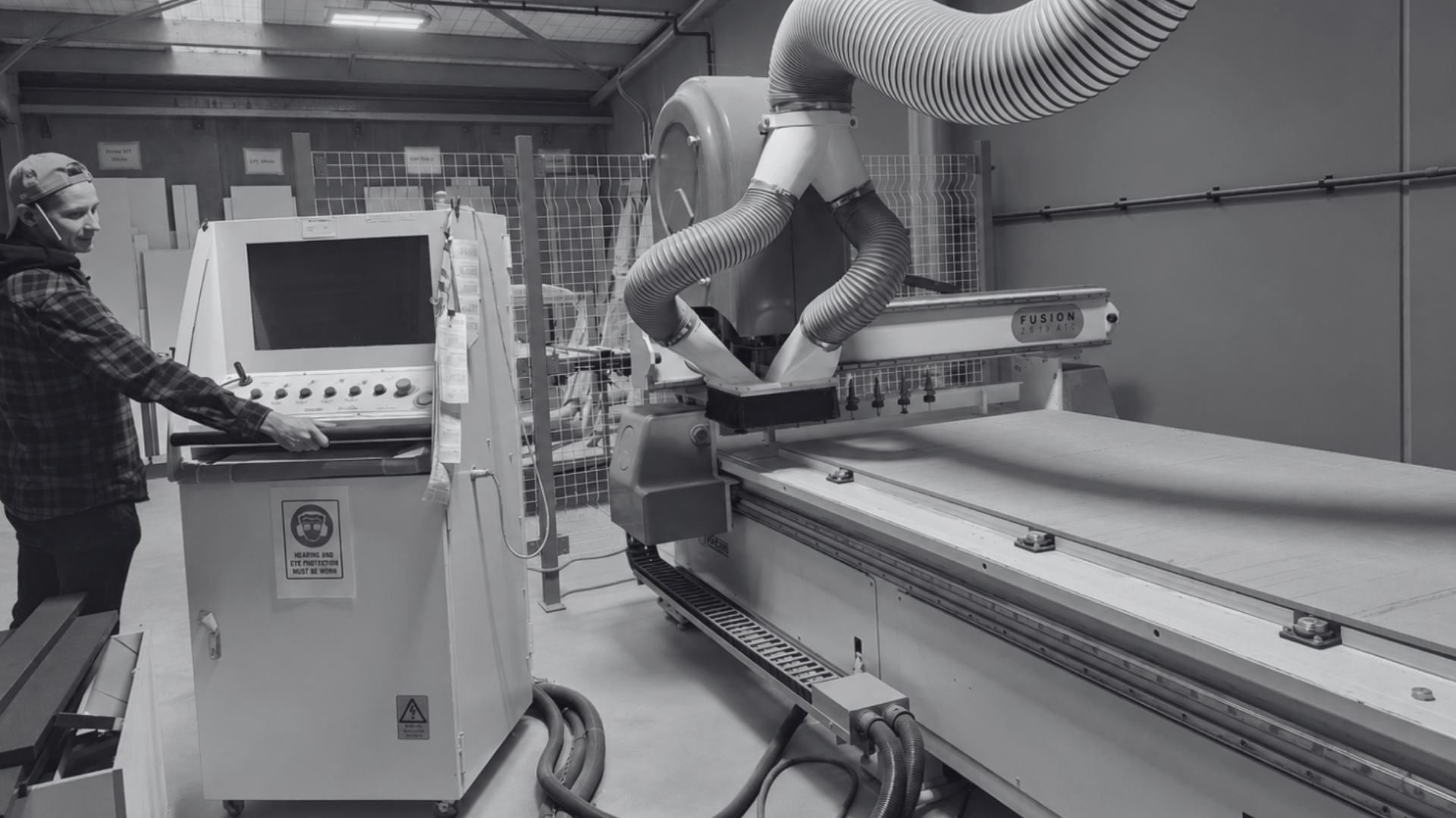 A person operating a large CNC machine in an industrial workshop. The machine has a control panel, large hoses for ventilation, and a flat working surface.
