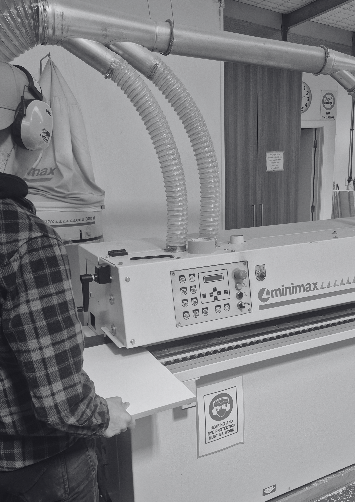 An industrial setting with a person working near a machine labeled 'minimax'. The person is wearing a plaid shirt and safety glasses, with a sign on the machine indicating the requirement to wear hearing and eye protection. There are large metal pipes and a cabinet in the background.