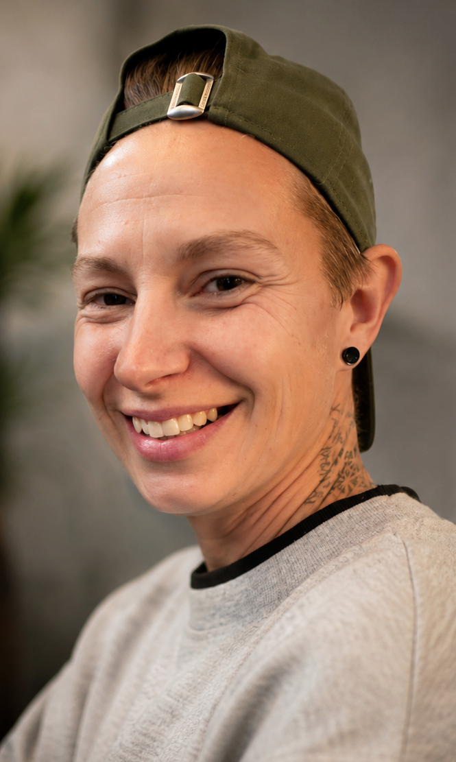 Close-up of smiling person wearing a green baseball cap worn backwards, black earrings, and a gray shirt with a black collar.