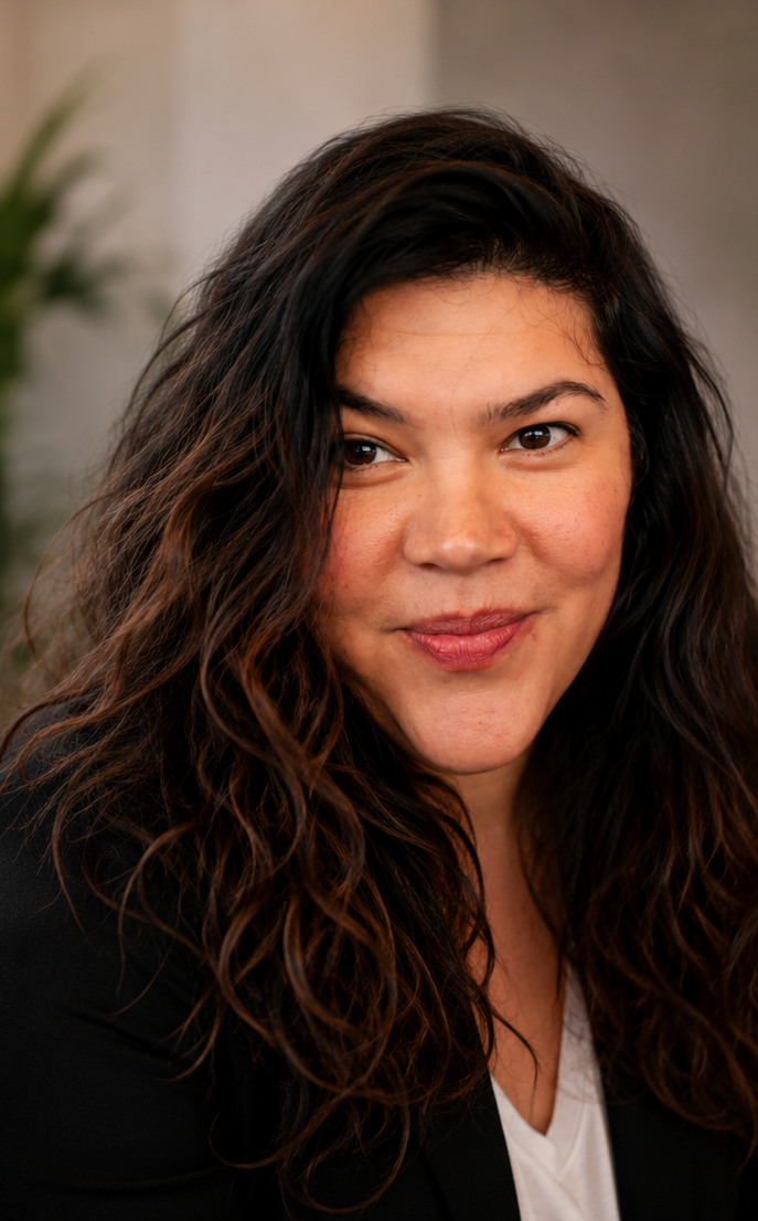 A woman with long, curly dark hair and light skin, smiling slightly, in an indoor setting with a blurred background.