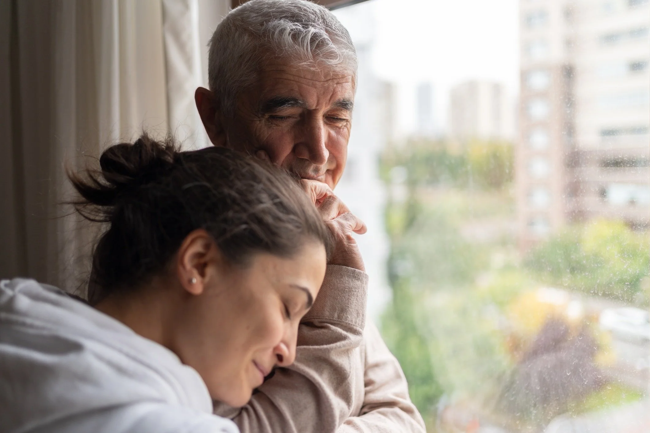 A man with gray hair and a woman with dark hair are resting their heads against each other while looking out a window.