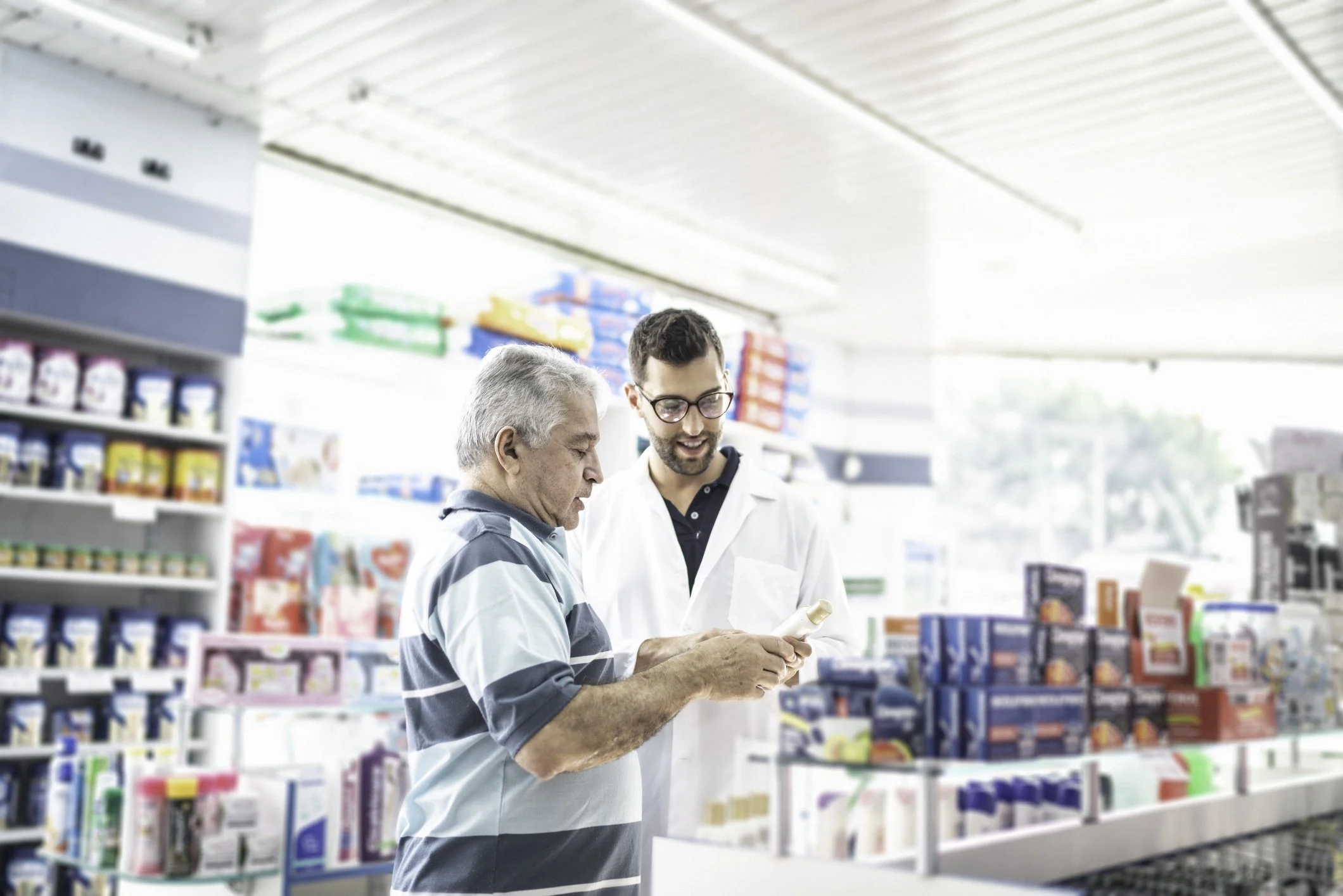 Two men, one older and one younger, are shopping in a pharmacy or drugstore aisle. The younger man in a white coat, possibly a pharmacist, is showing something to the older man in a checkered shirt. Shelves around them are stocked with various health and beauty products.