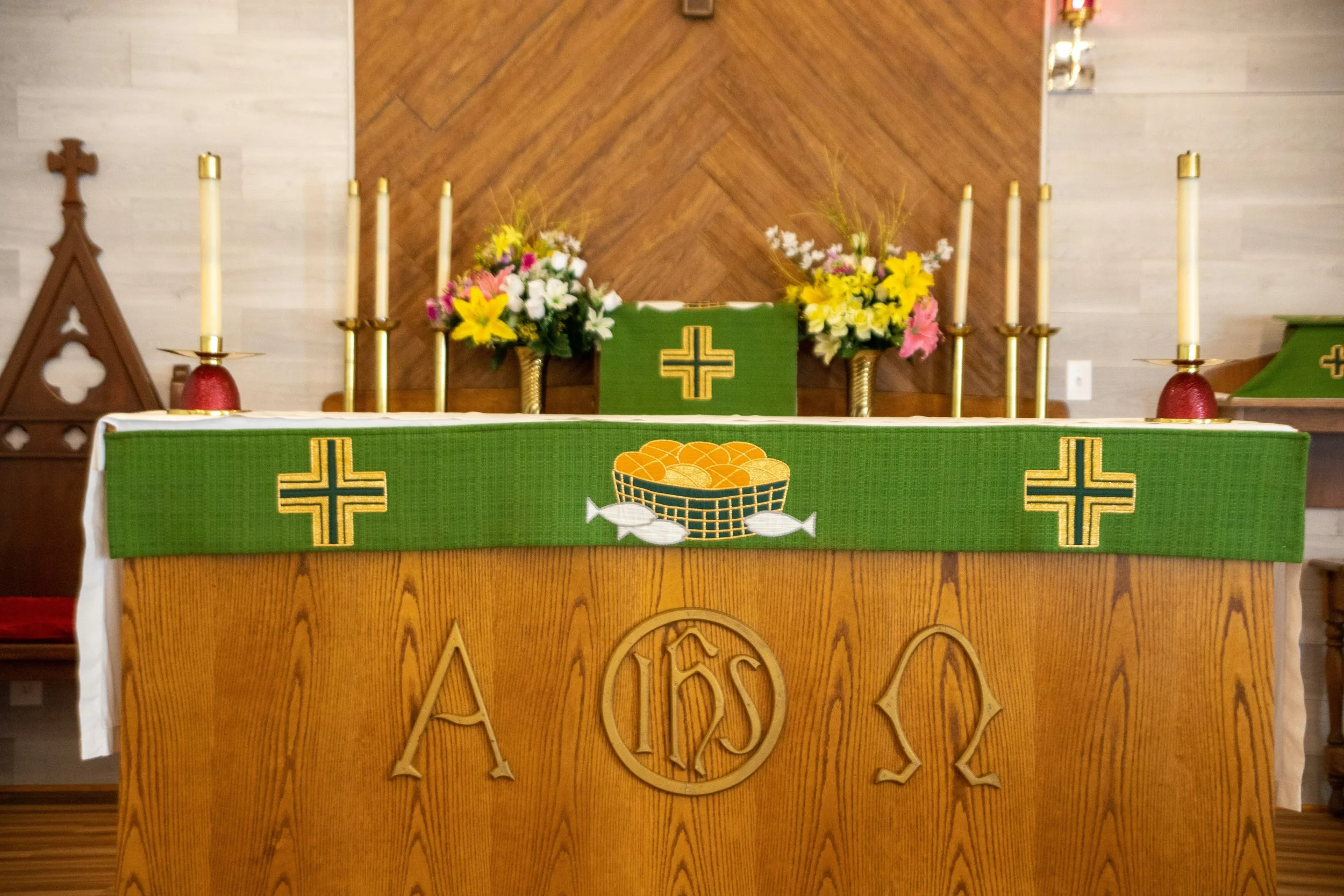An altar decorated with green cloth featuring yellow crosses and a basket of bread and fish symbols. On top are two flower arrangements with pink, yellow, and white flowers, and several white candles in gold holders. The background has a wood paneled wall with a cross on the left side.