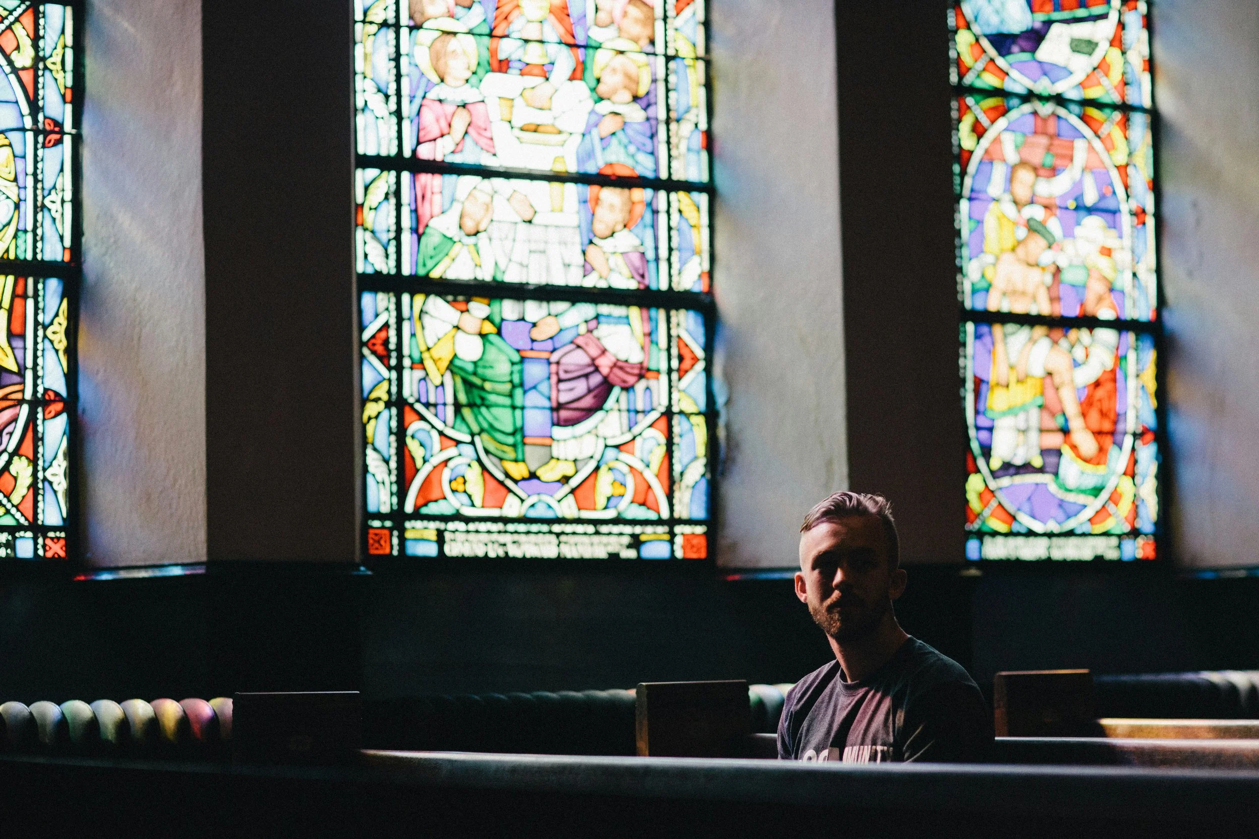 A man sitting in front of a row of colorful bowling balls with stained glass windows featuring biblical scenes in the background.