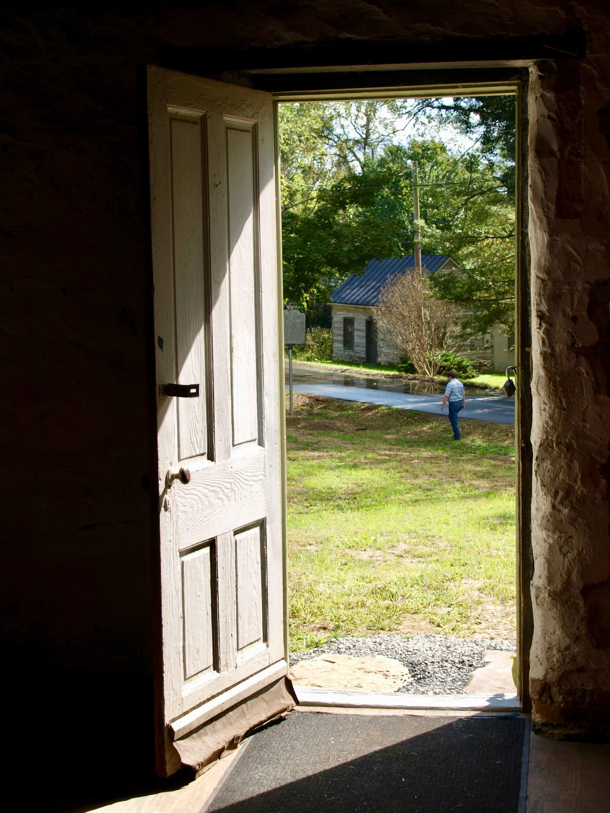 Open wooden door leading outdoors to a sunny scene with trees, a person walking, and a small building with a metal roof.
