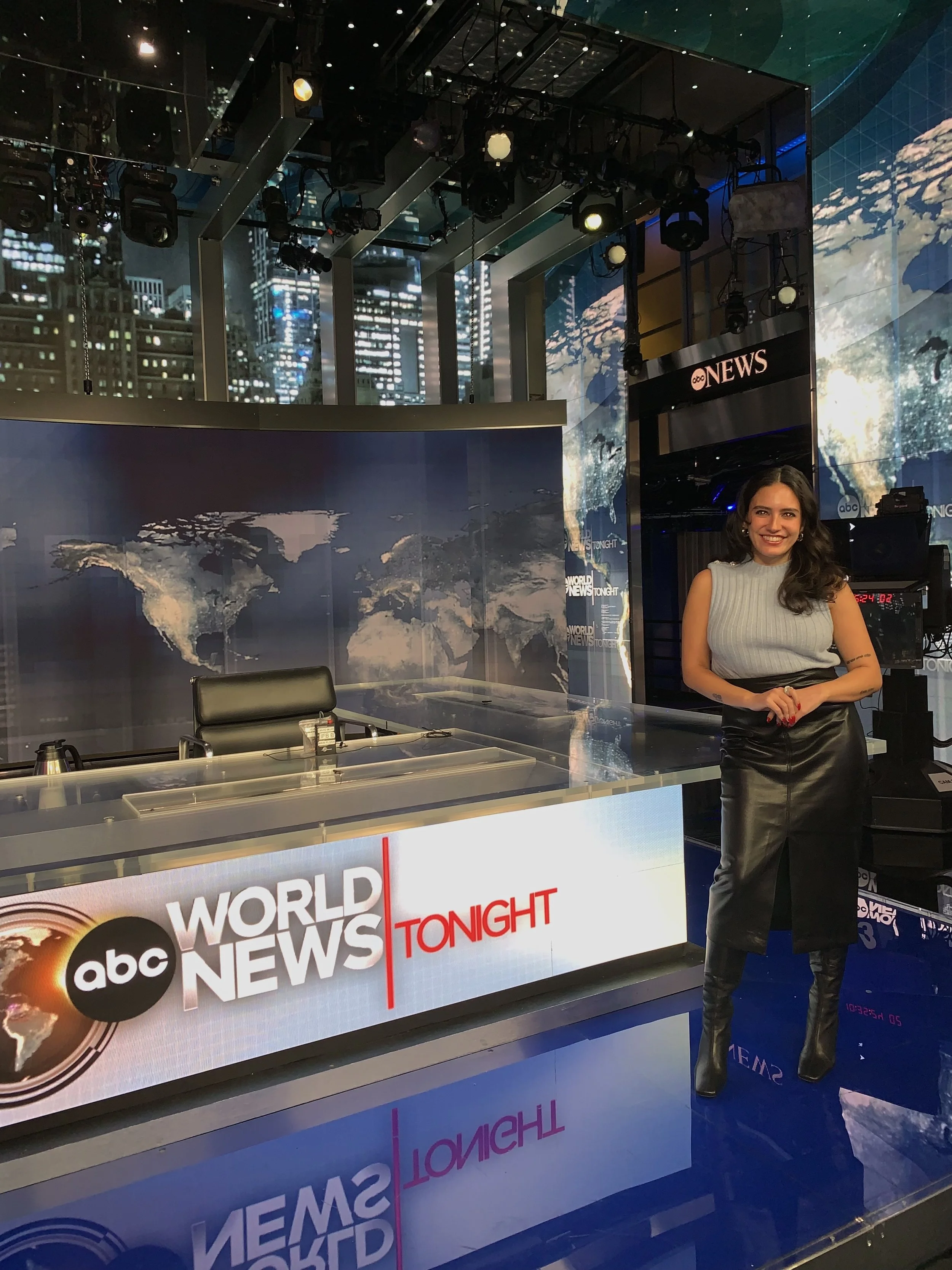 A woman standing in a television news studio with ABC News and World News Tonight logos on the front desk, a large digital world map on the background, and studio lighting equipment hanging from the ceiling.