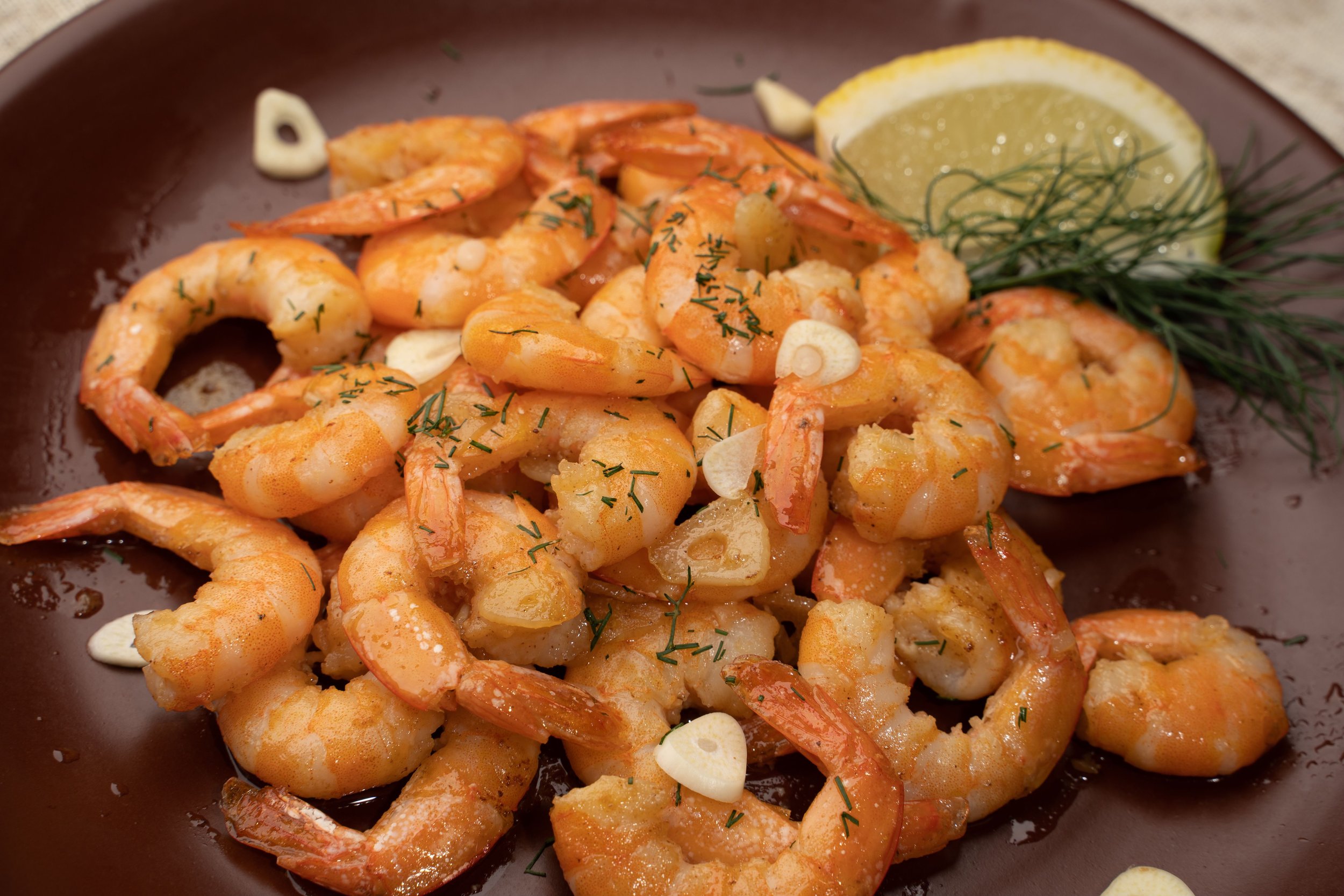 Close-up of a plate of cooked shrimp garnished with chopped herbs, garlic slices, a lemon wedge, and fresh dill.