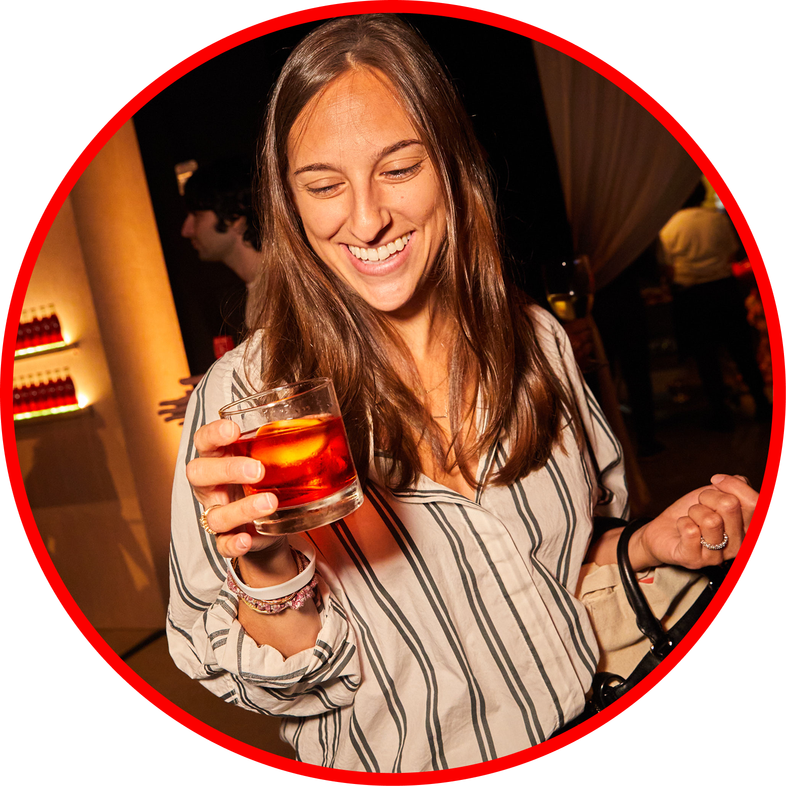 A woman smiling and holding a glass of red drink with ice on a night out in a bar or club setting.