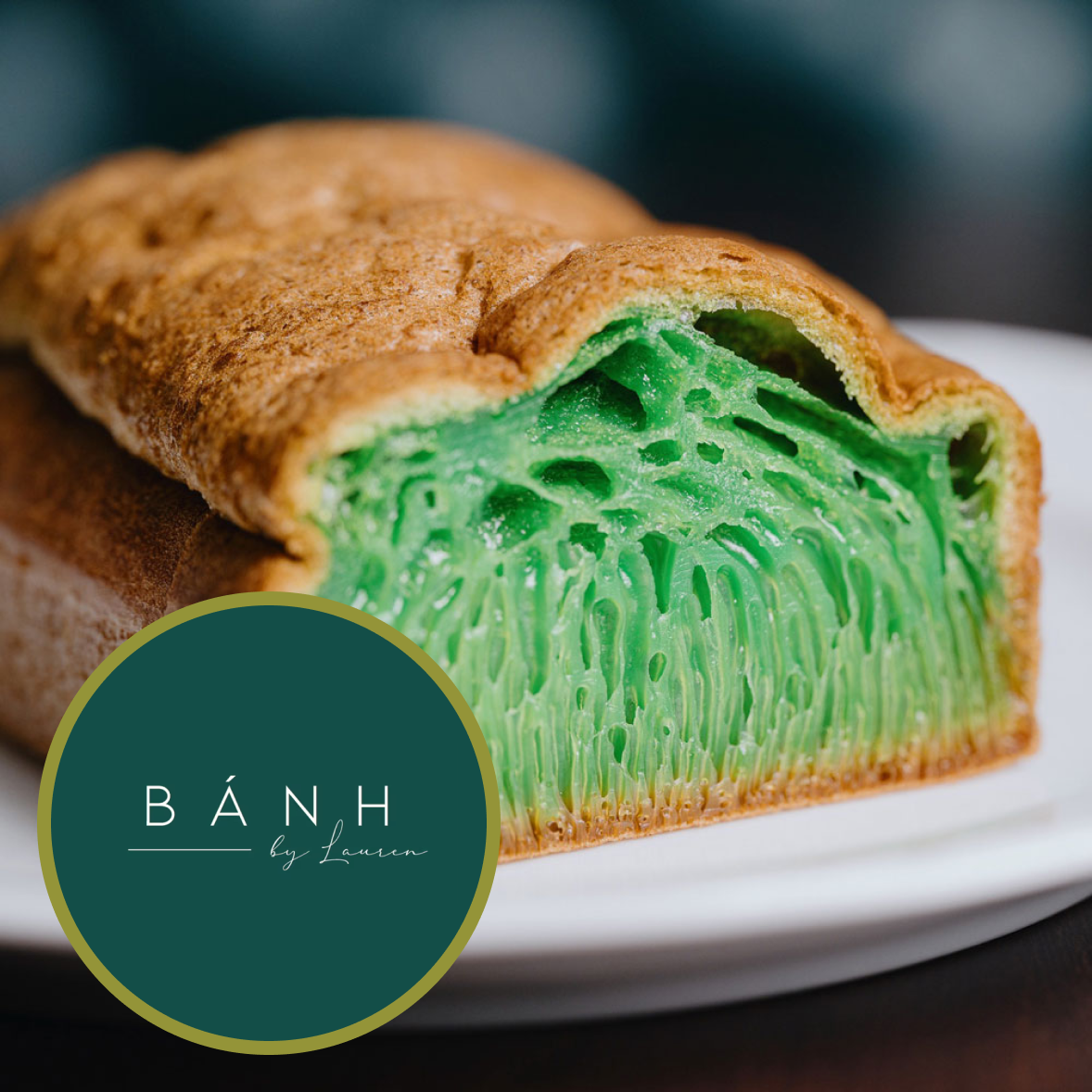 Close-up of a green, jelly-like pastry with a golden-brown crust on a white plate. The pastry has a detailed, web-like interior texture. Logo overlay with the text 'BÁNH by Lauren' in a dark green circle.