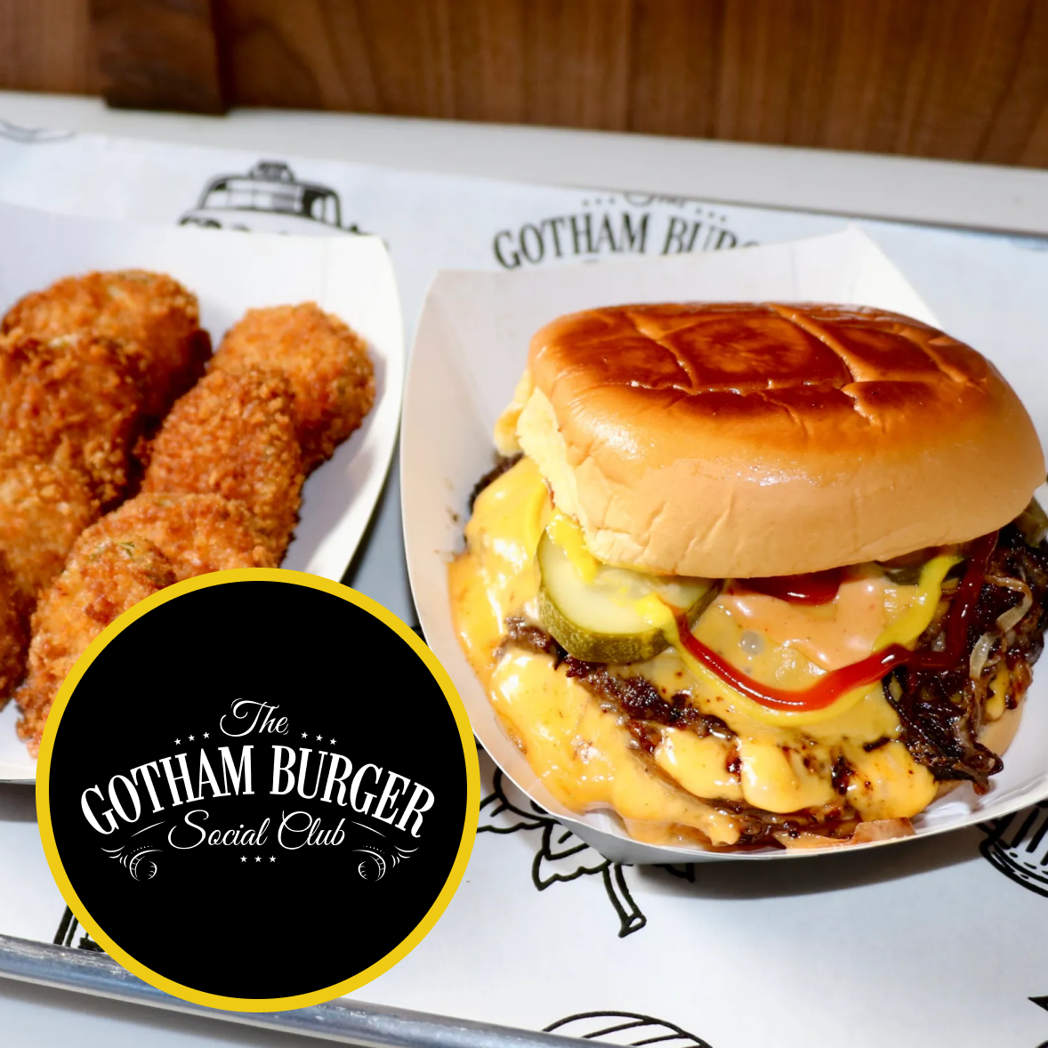 Tray of food including a fried chicken strip, cheeseburger with pickles, a side of French fries with ketchup, and a loaded nacho-style dish with cheese, jalapeños, tomatoes, and sour cream on a wooden table. A drink with a straw and a logo reading 'The Gotham Burger Social Club' is also visible.