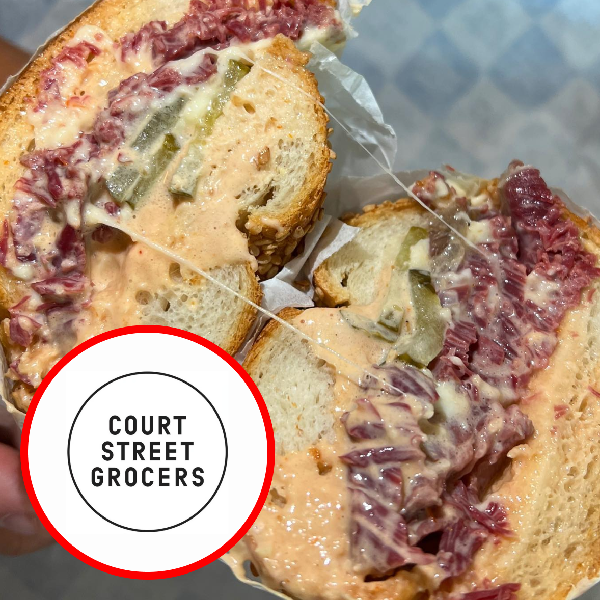Sandwich with sliced meat, greens, and melted cheese on focaccia bread, with a logo for Court Street Grocers.