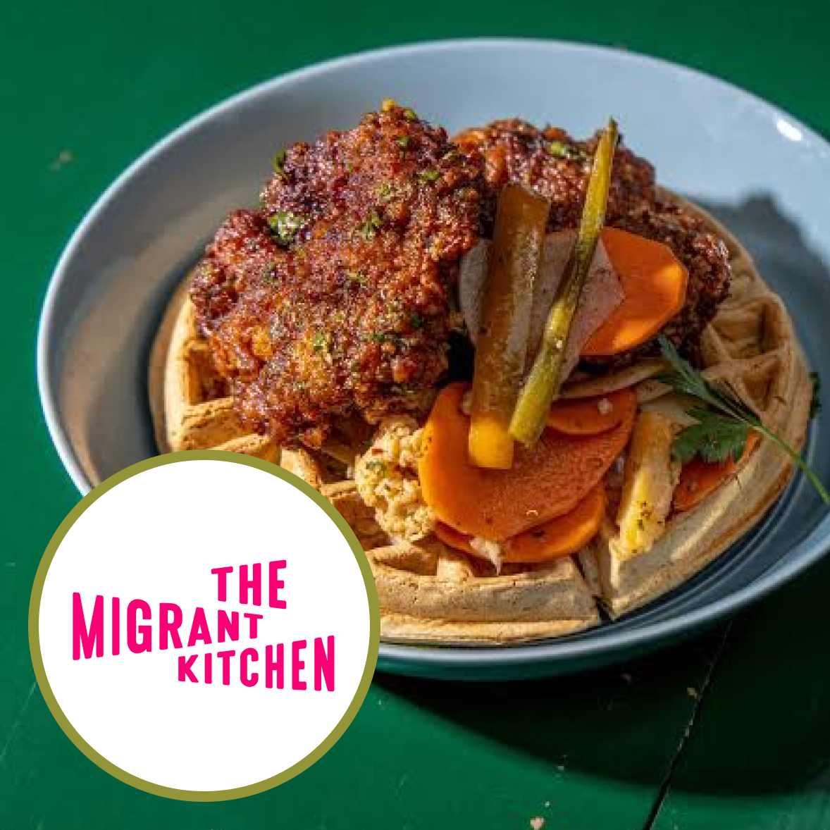 Plate of cooked meat, vegetables, and noodles with a 'The Migrant Kitchen' logo in the bottom left corner.