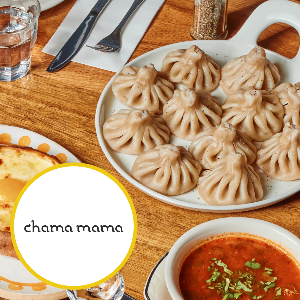 Plate of khinkali dumplings on a white dish, glass of water, bowl of tomato-based soup garnished with herbs, and utensils on a wooden table.