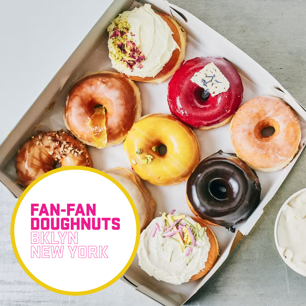 A box of assorted colorful donuts topped with frosting and sprinkles on white paper, with text overlay reading 'Fan-Fan Doughnuts Brooklyn New York'.