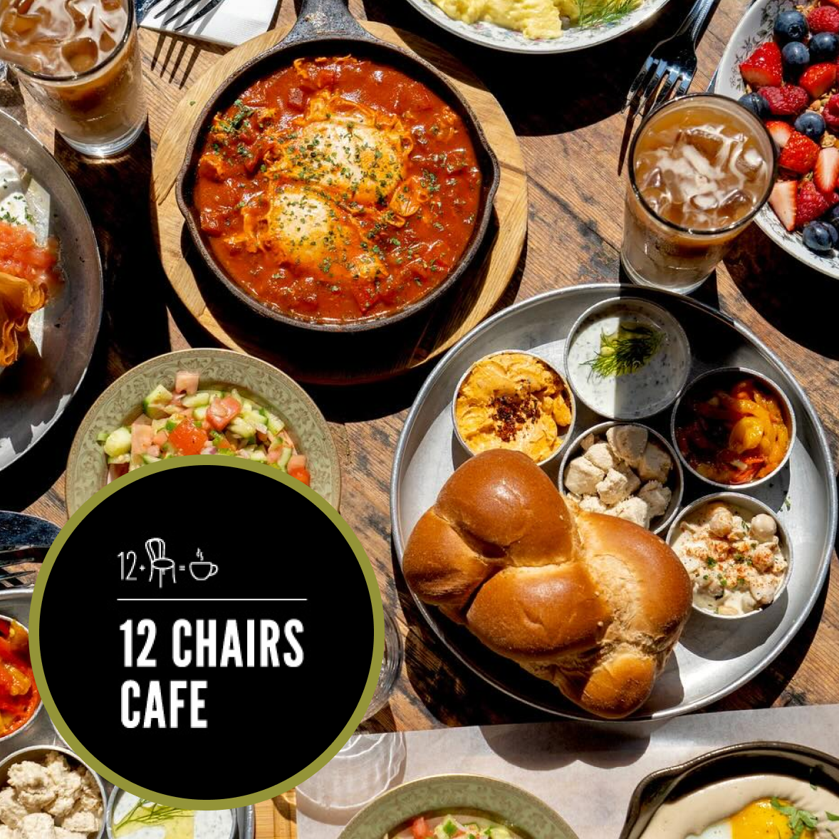 An overhead shot of a table with various breakfast dishes, including a skillet of eggs in tomato sauce, a bread roll, bowls of yogurt and fruit, a salad, and glasses of iced drinks. There is a black circular graphic with white text that reads '12 Chairs Cafe' and icons of a chair, a hat, and a coffee cup.