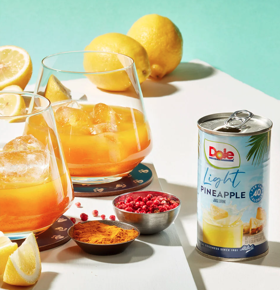 Box of Dole fruit bowls with three individual bowls of fruit salad on a wooden surface, three gold spoons, and a colorful fruit-themed package in the background.
