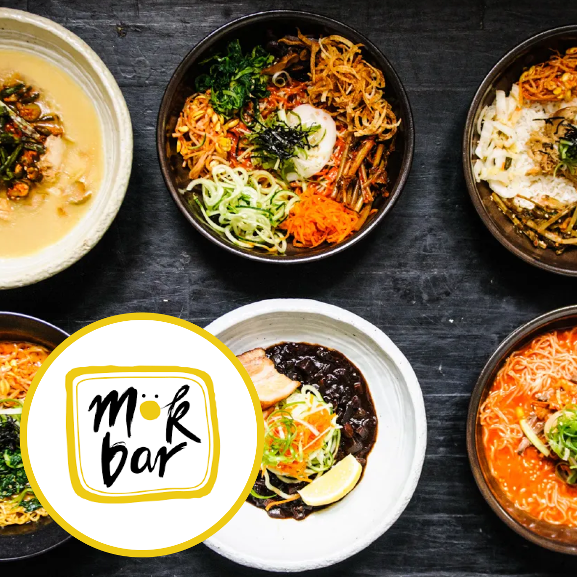 Korean dishes including bibimbap in bowls with vegetables, rice, and sauces, with a scoop of black bean paste and a lemon wedge, and a logo for 'Mork Bar' on the overlay.