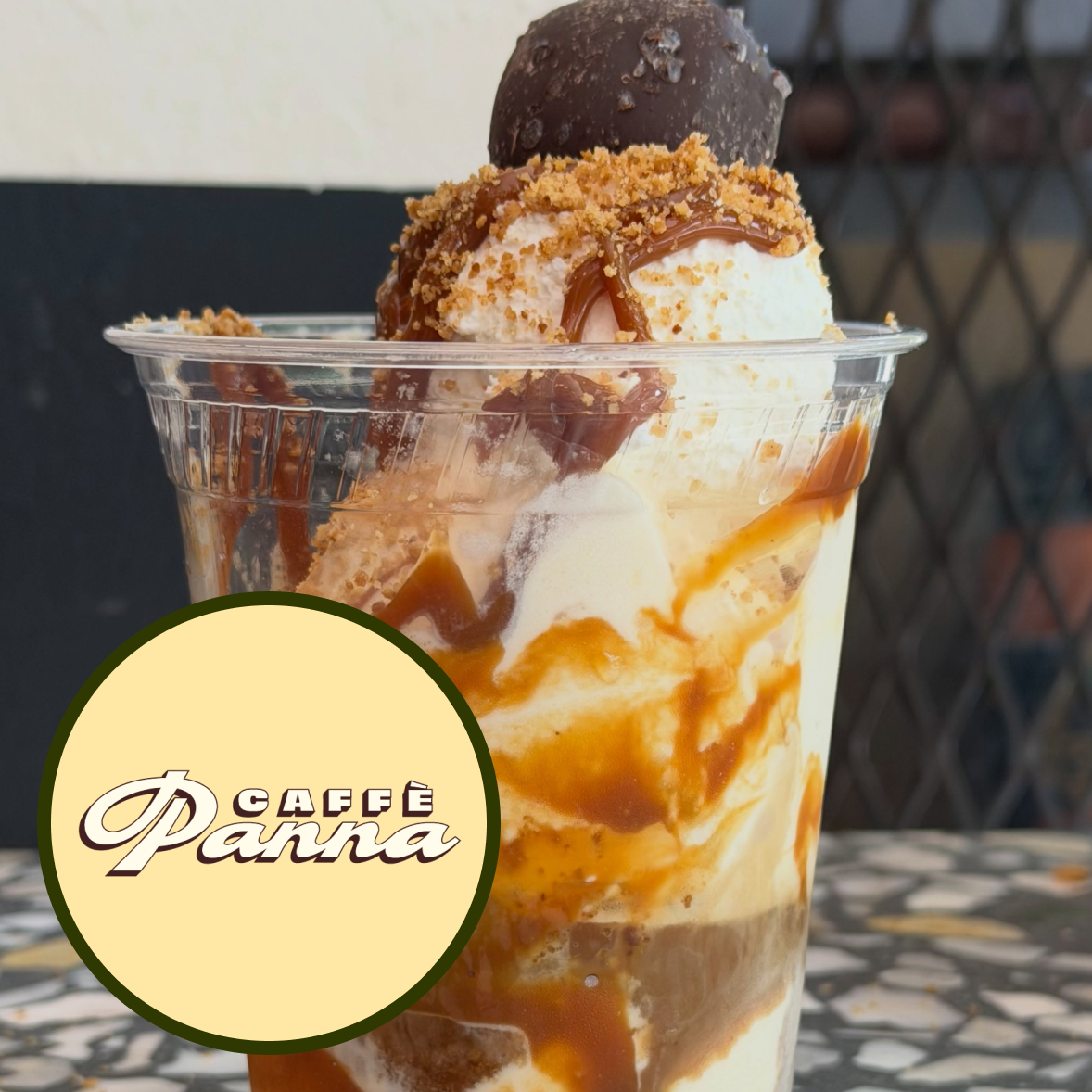 A glass of affogato topped with whipped cream, chocolate shavings, caramel sauce, and sesame seeds with a bottle of amaretto in the background and a logo for Café Panna in the front.