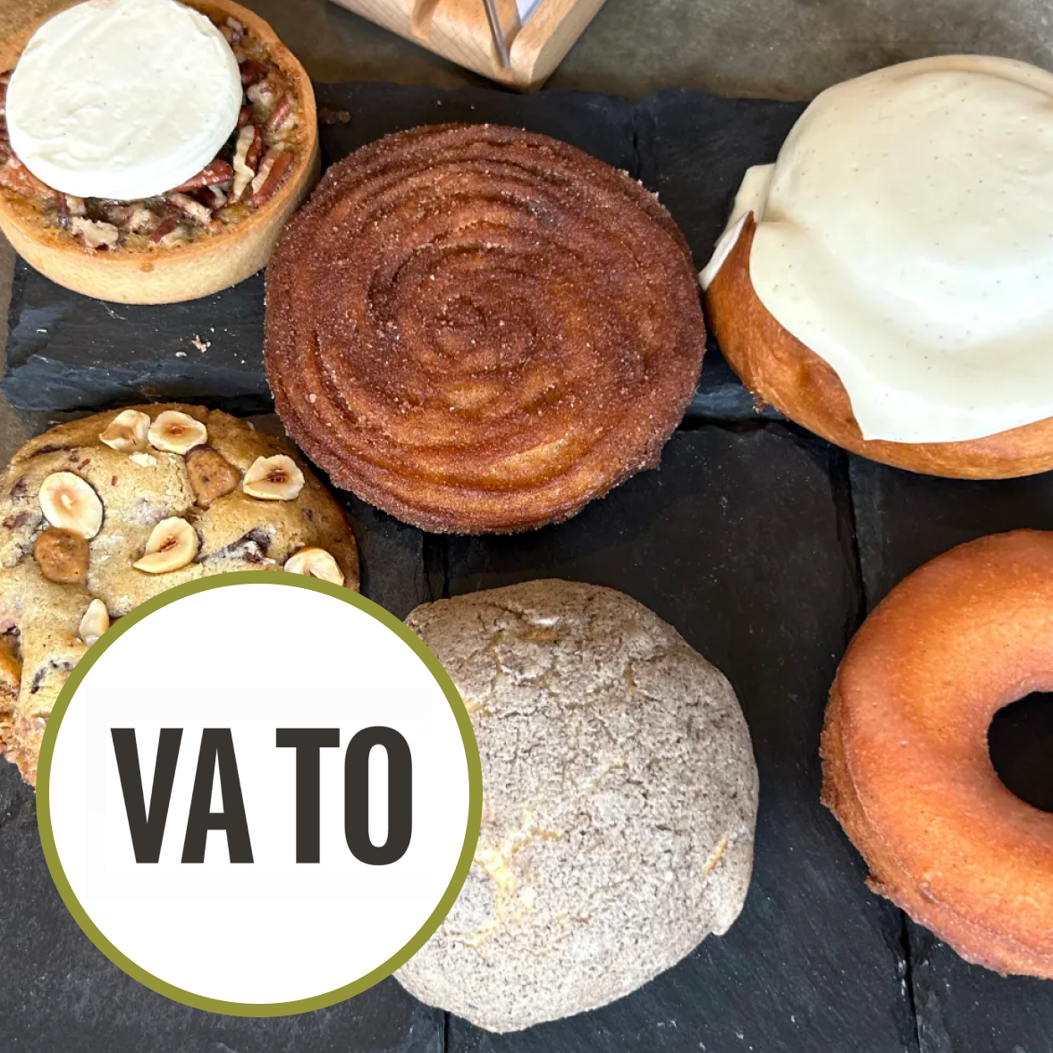 Variety of baked goods on black slate board, including a cinnamon roll, a donut, a cookie topped with hazelnuts, and a cream-filled pastry with white icing.