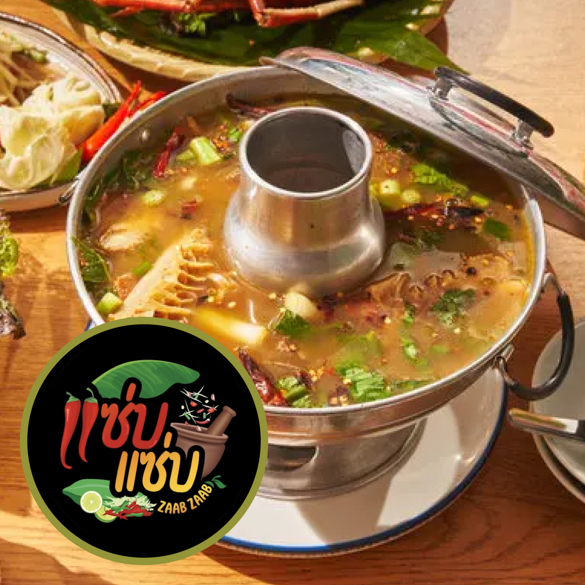 A spicy Thai hot pot with herbs and vegetables, surrounded by fresh ingredients like lemon and chili peppers, on a wooden table with a Zaab Zaab logo.