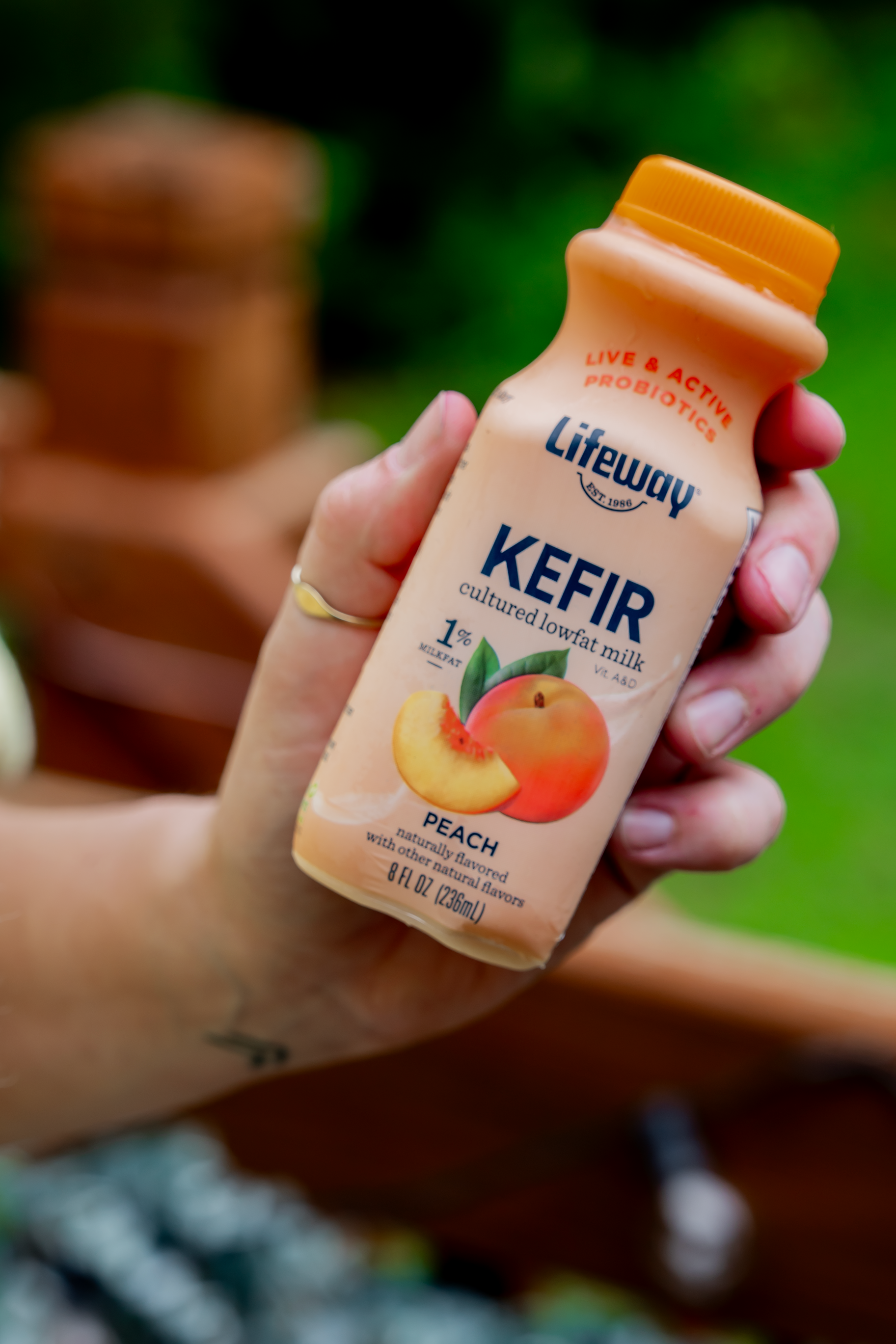 A hand holding a carton of Lifeway Kefir peach flavored probiotic cultured lowfat milk with a blurred background of green outdoors.