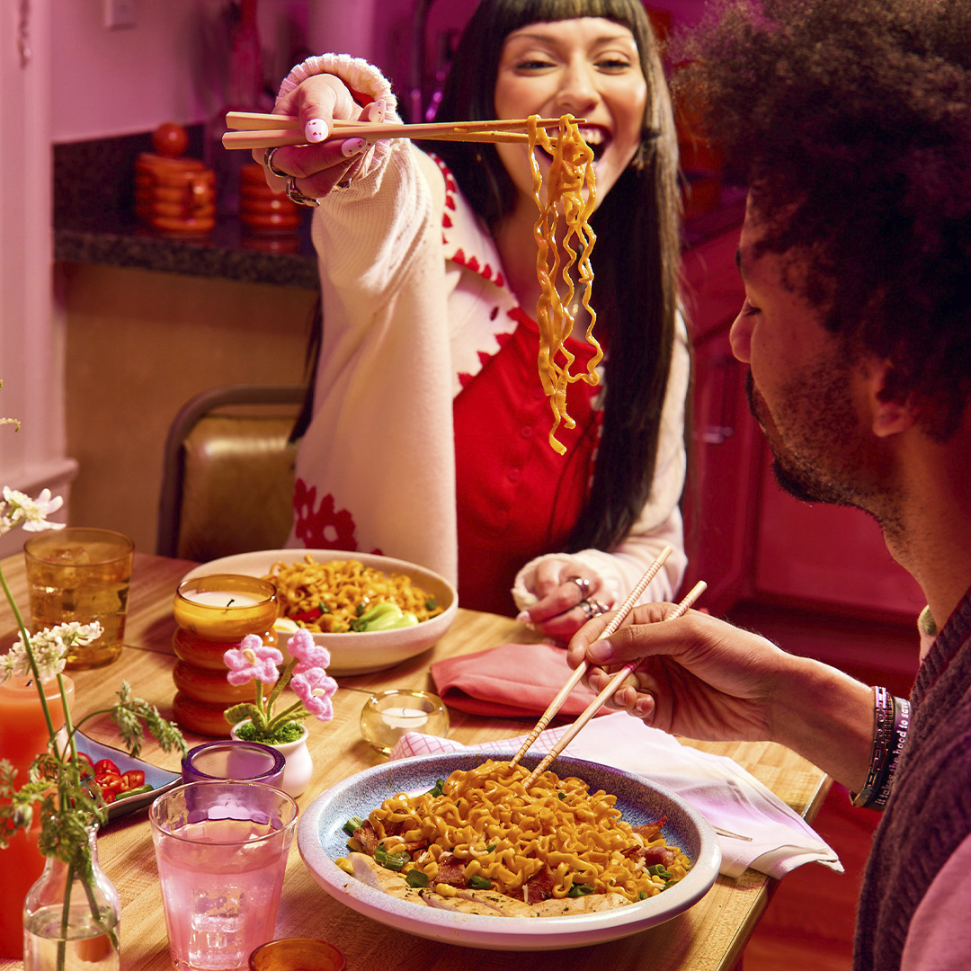 A bowl of ramen noodle soup topped with chopped green onions and fried chicken, surrounded by four Maruchan ramen noodle packages on a green surface with white sports lines.