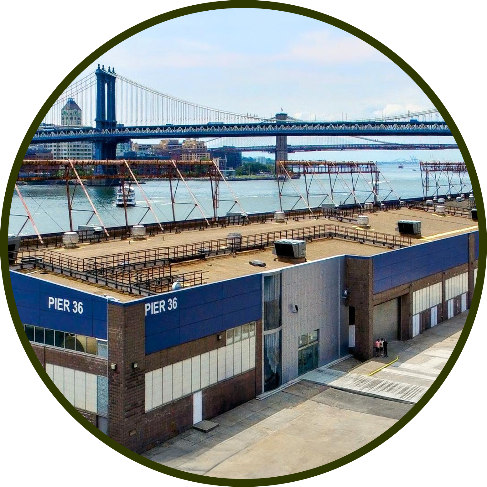 View of Pier 36 with a blue exterior and multiple large windows, located along the waterfront with the New York City skyline and the Manhattan Bridge in the background.