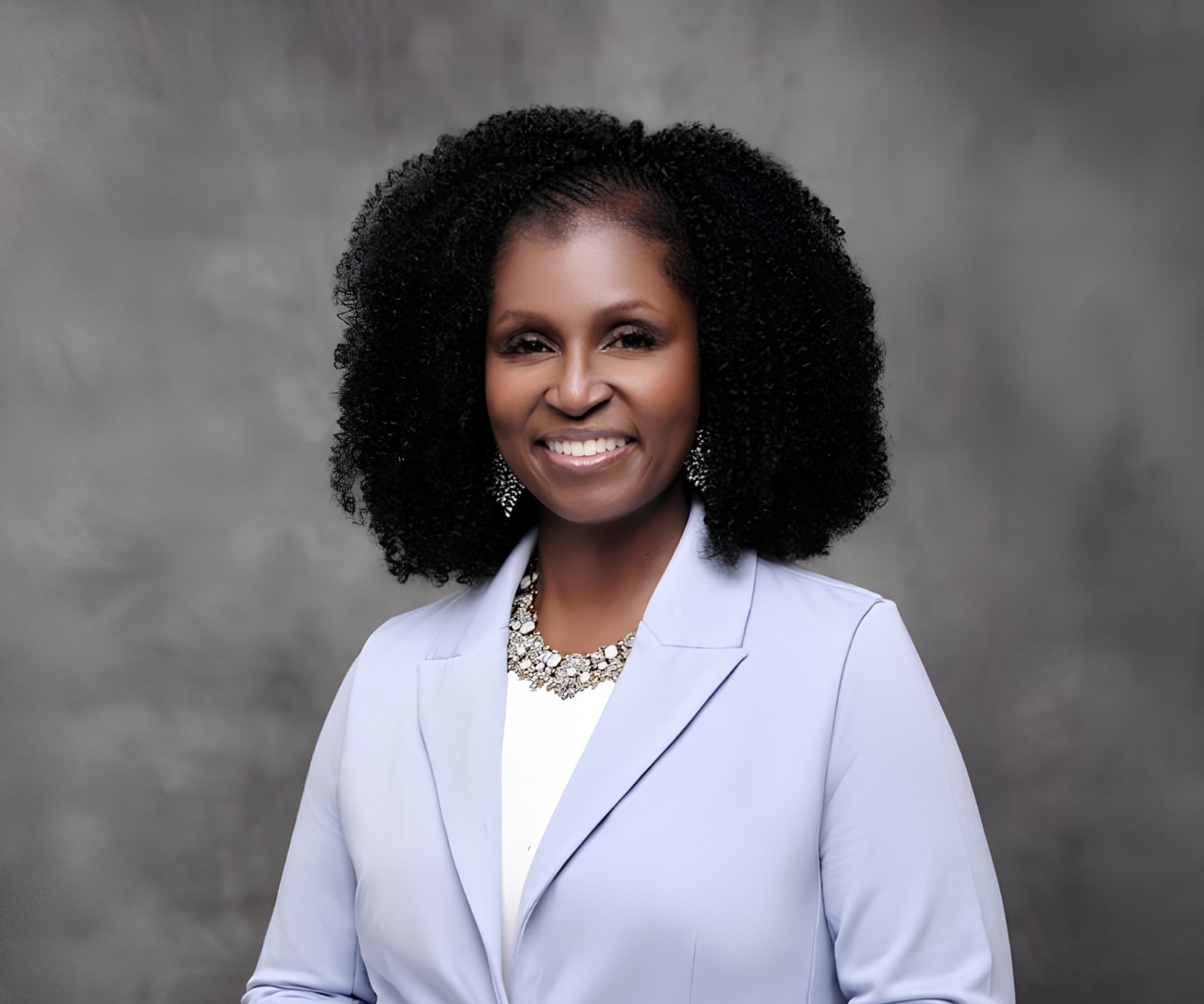 Portrait of an African American woman smiling, wearing a light gray blazer, white top, and a sparkly statement necklace, against a gray textured background.