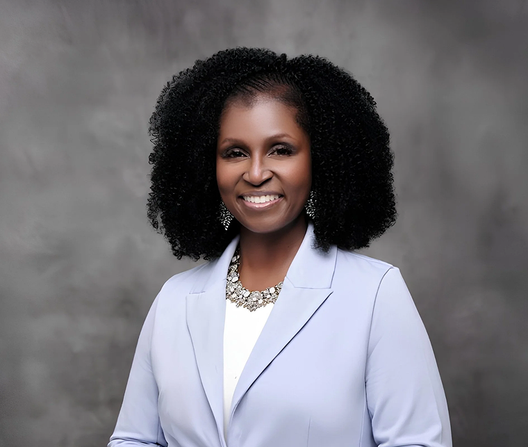Portrait of a smiling Black woman with curly black hair, wearing a white blazer, a statement necklace, and earrings against a gray background.