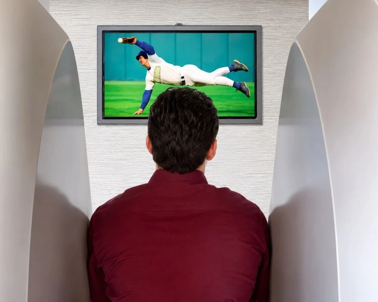 A man sitting in a viewing pod watching a TV screen that shows a baseball player catching a fly ball.