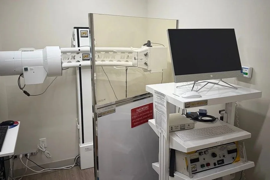 Medical imaging room with a large machine connected to a transparent shield, a computer monitor on a white cart, and various medical equipment.