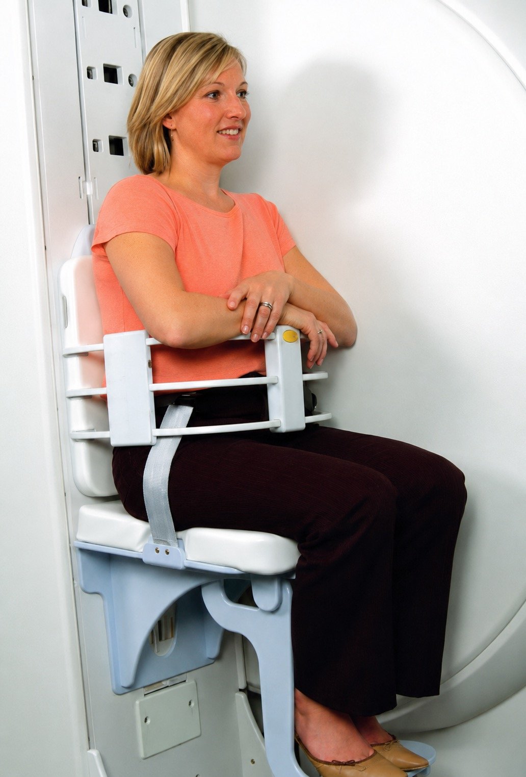 A woman sitting in a seat with a safety harness installed, inside an airplane or medical transport vehicle, with a white background and wall panels.