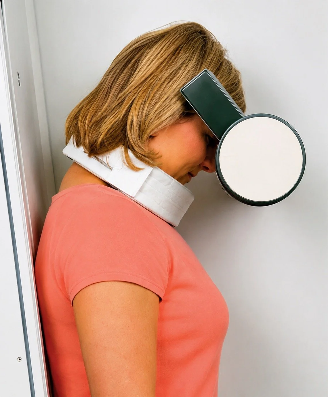 A woman with short blonde hair in a peach-colored shirt is standing with her head against a white wall. She is wearing a special cervical collar and a head-bearing device for medical or therapy purposes.