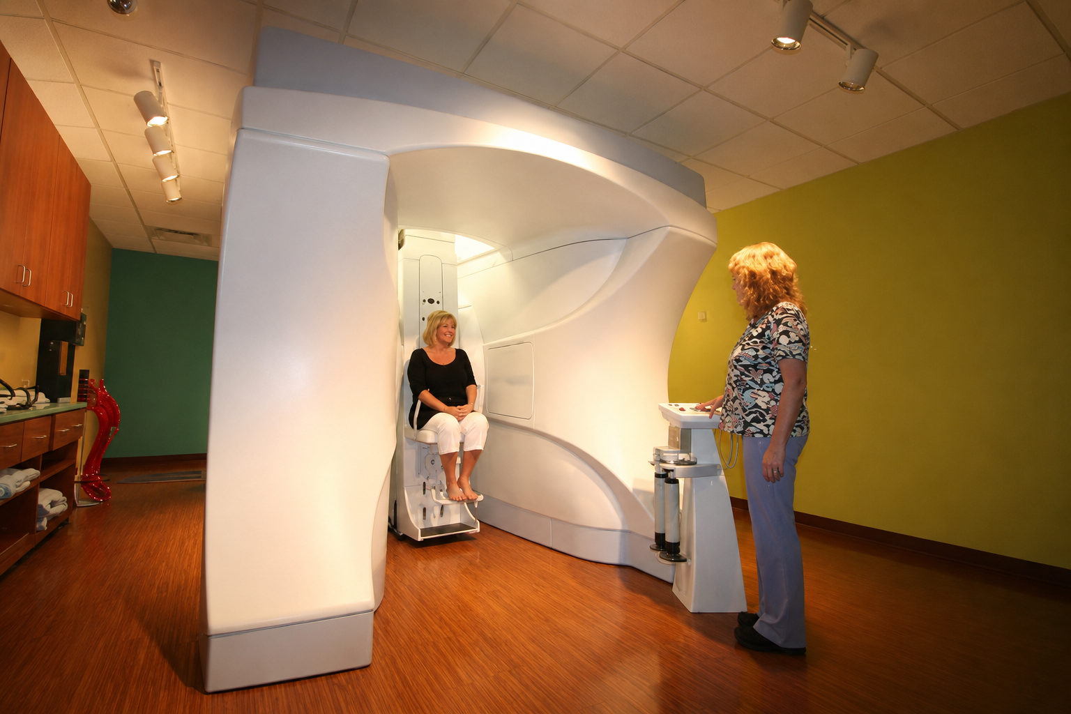 Woman sitting inside an MRI machine while a technician stands nearby operating controls in a medical facility.