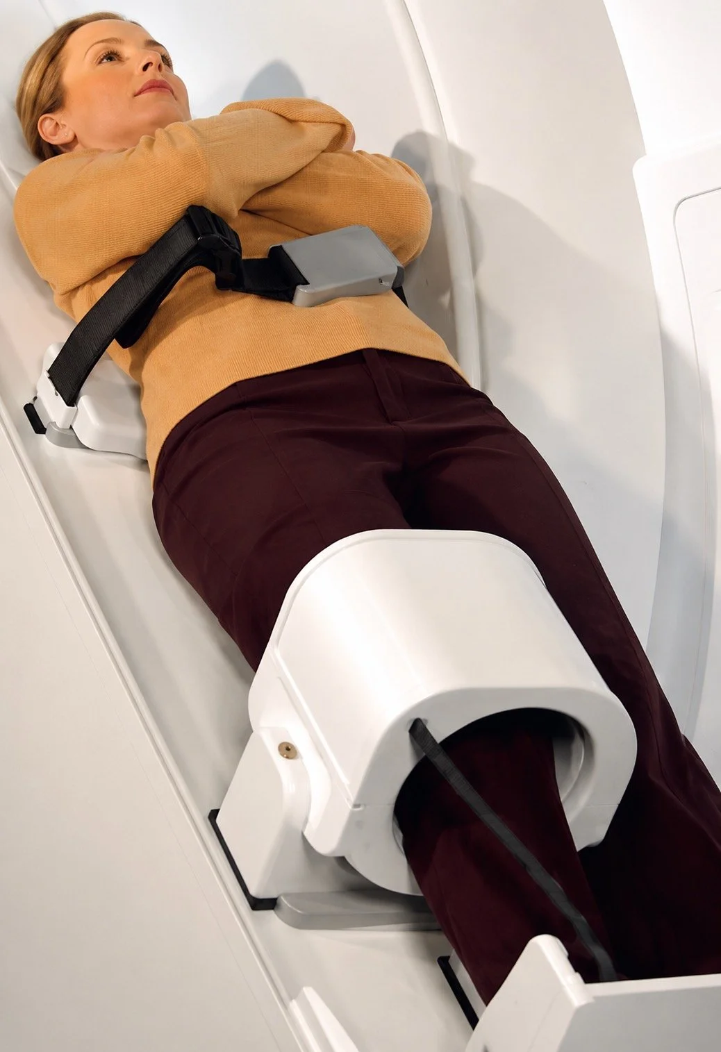 A woman lying on a medical imaging table with her legs inside an MRI machine, wearing a beige sweater and red pants.