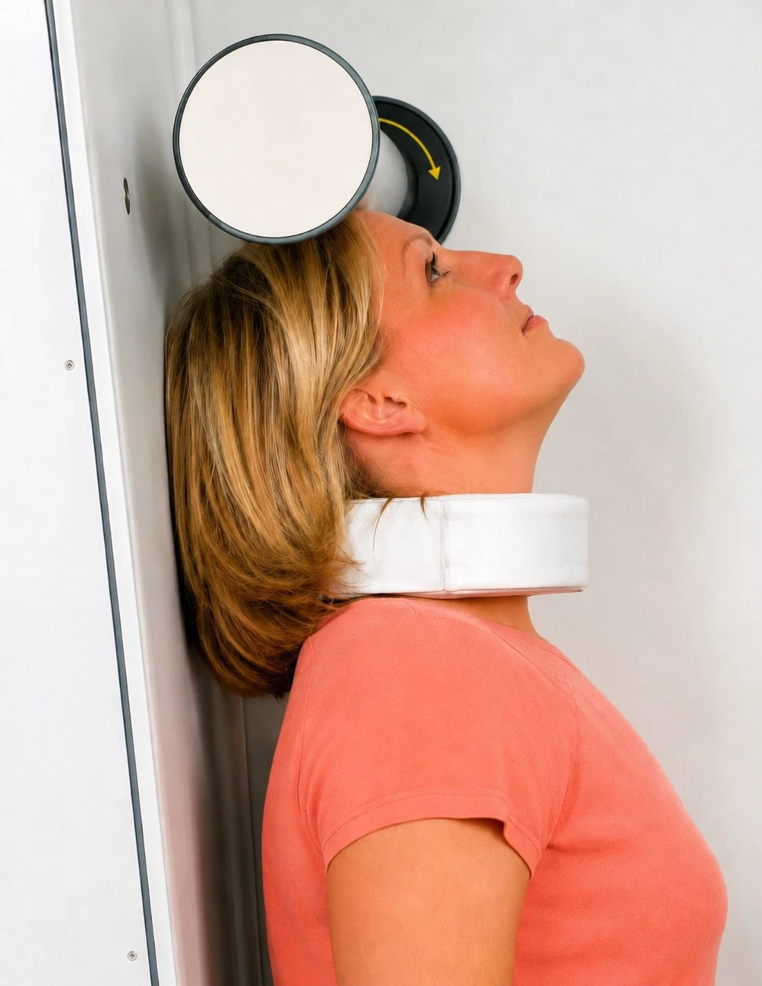 Woman undergoing a medical imaging scan, with her head resting on a supportive foam pad and her face positioned against a circular black and white device with a yellow arrow, in a clinical setting.